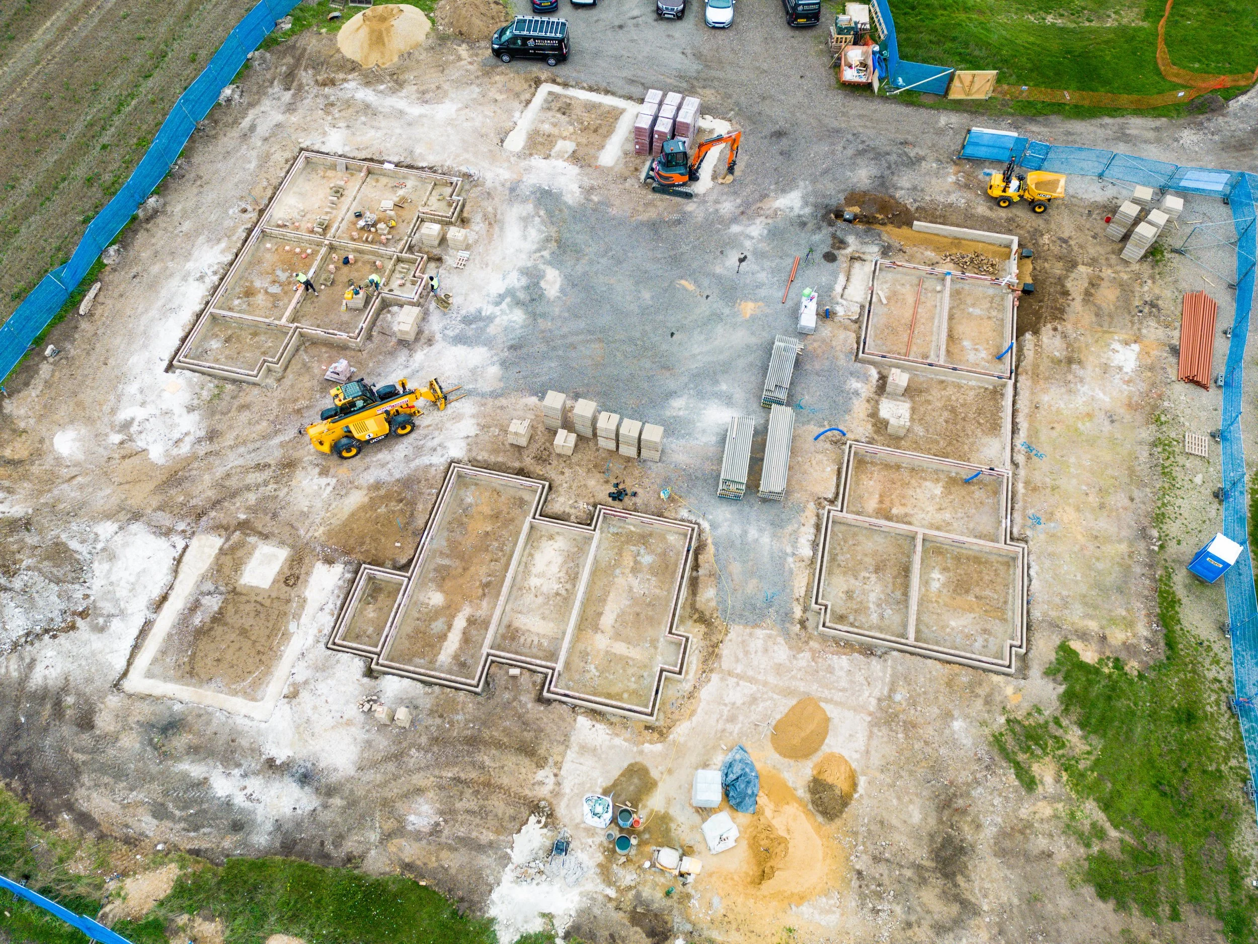 An aerial view of a construction site with foundation outlines, construction equipment including a yellow forklift, and vehicles parked at the edge of the site.