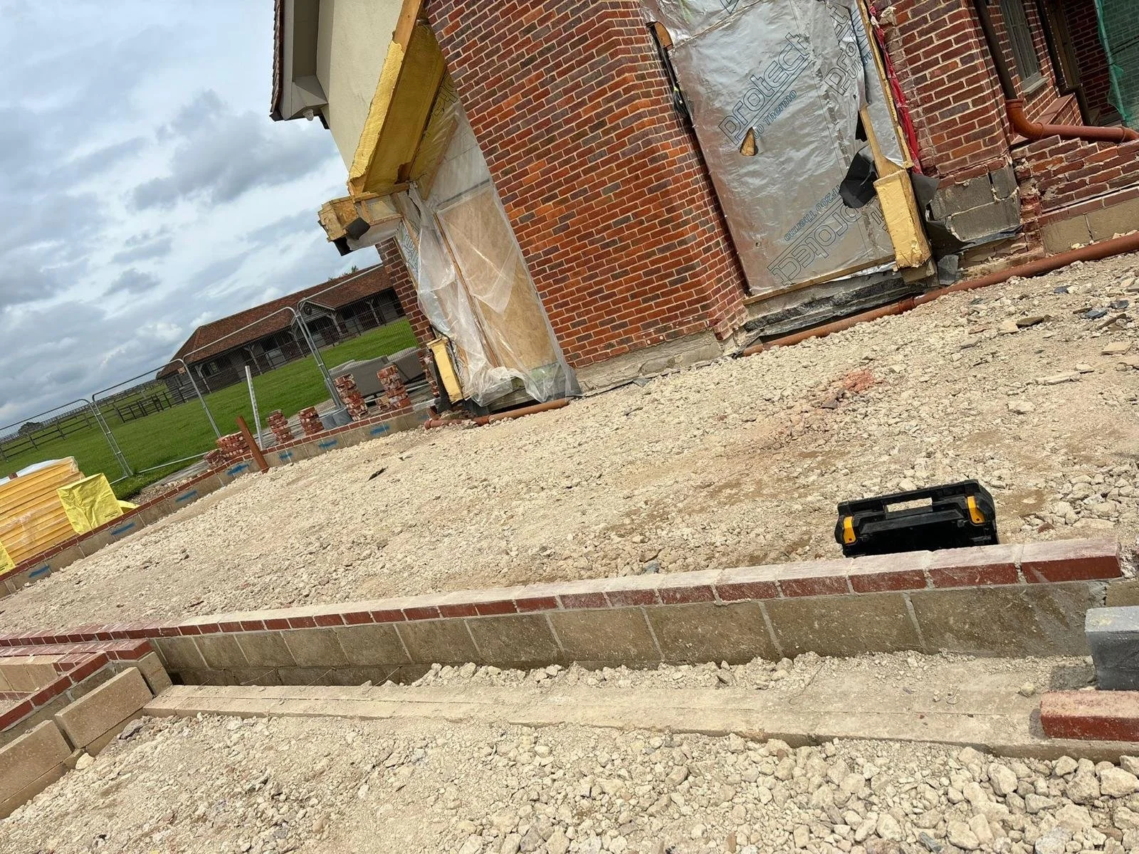 Construction site with a brick building in progress, gravel ground, orange pipes, and construction materials visible under a cloudy sky.