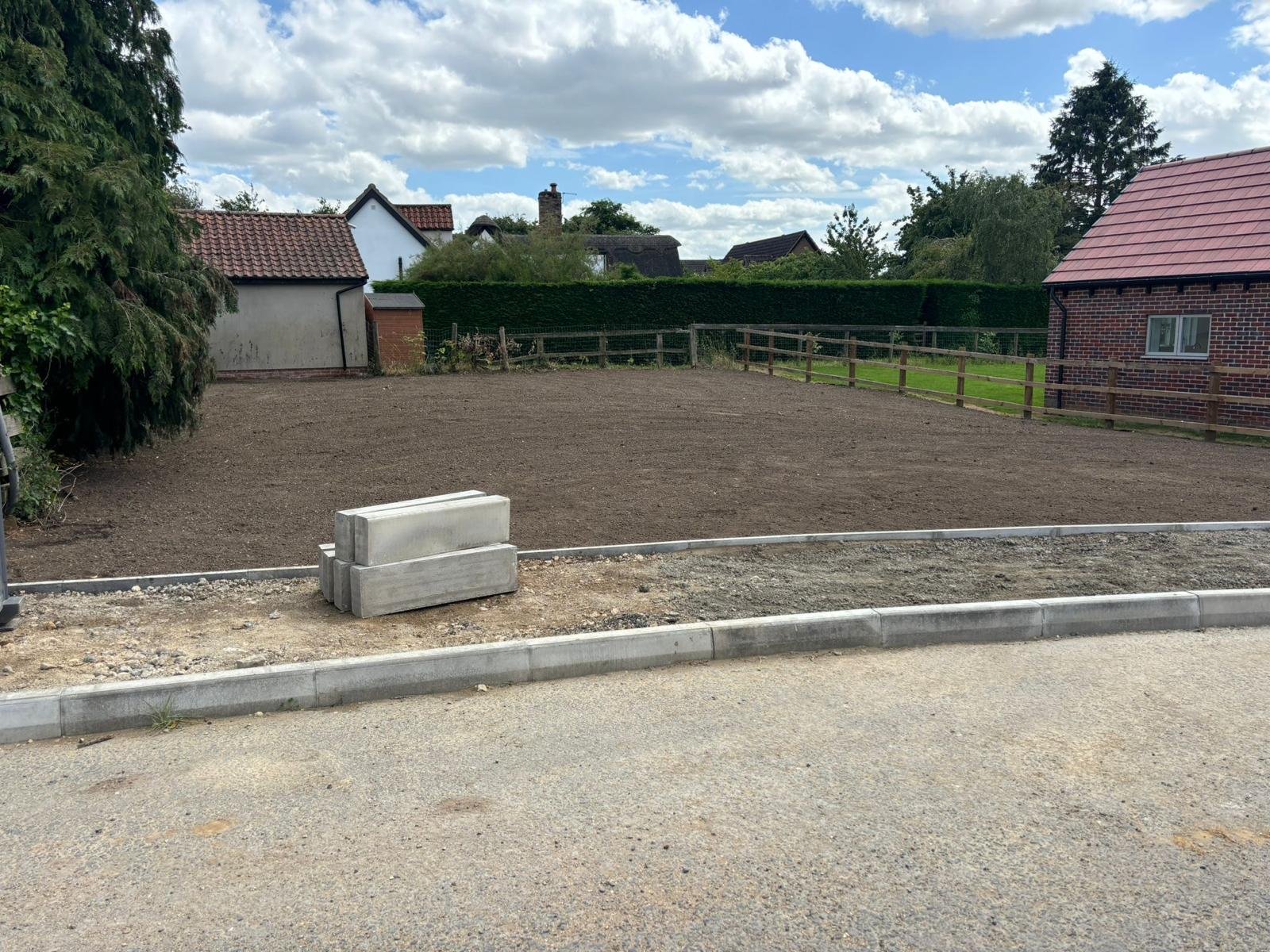 A vacant lot with a dirt surface, cordoned off from the road with concrete curbing and a pavement. In the foreground, there are two bricks placed on the curb. The background features trees, bushes, and a few houses with red-tiled roofs under a partly cloudy sky.