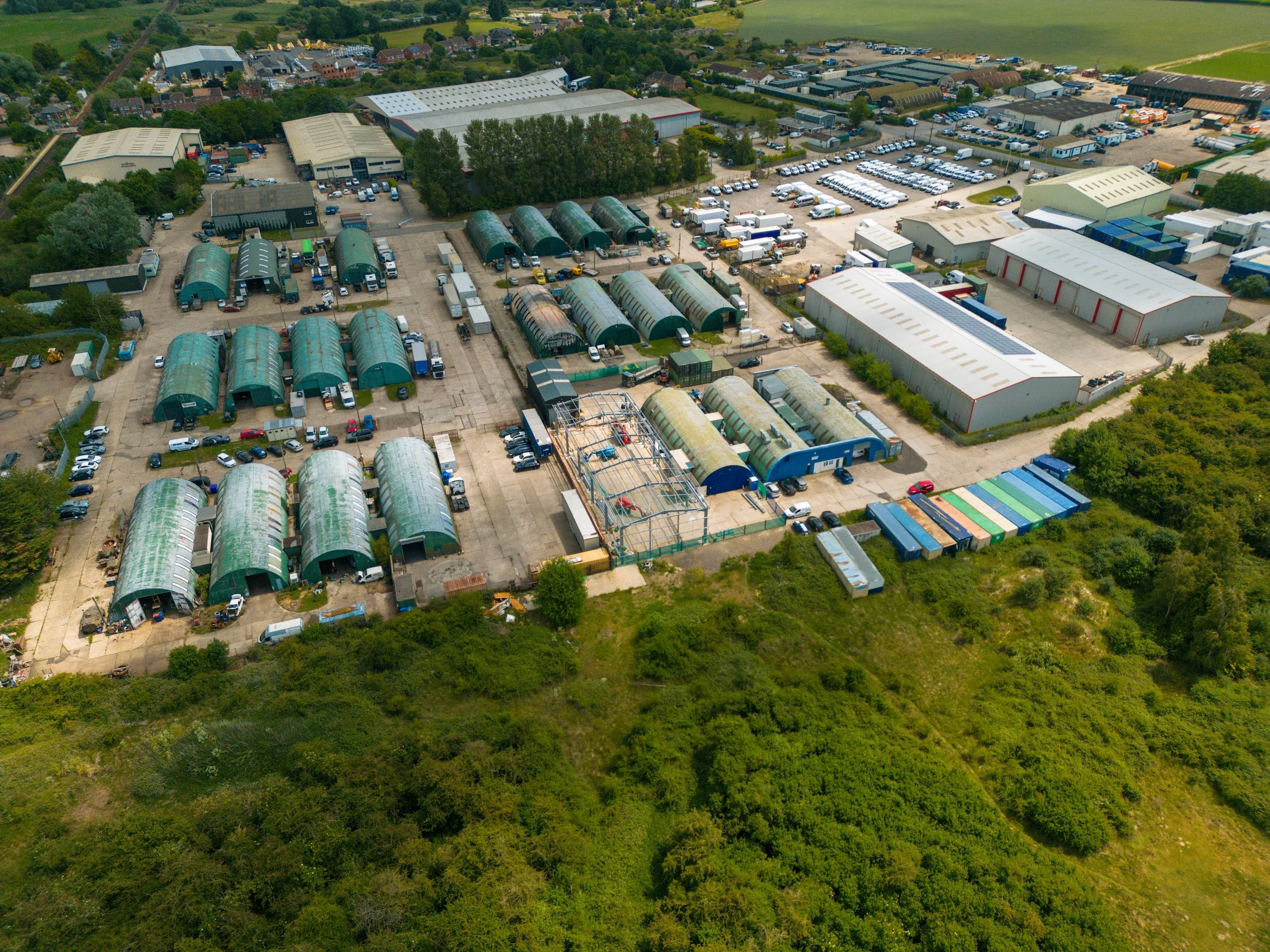 Aerial view of an industrial storage facility with numerous green and white warehouse buildings, greenhouses, and a large parking lot with many cars and trucks, surrounded by trees and green fields.