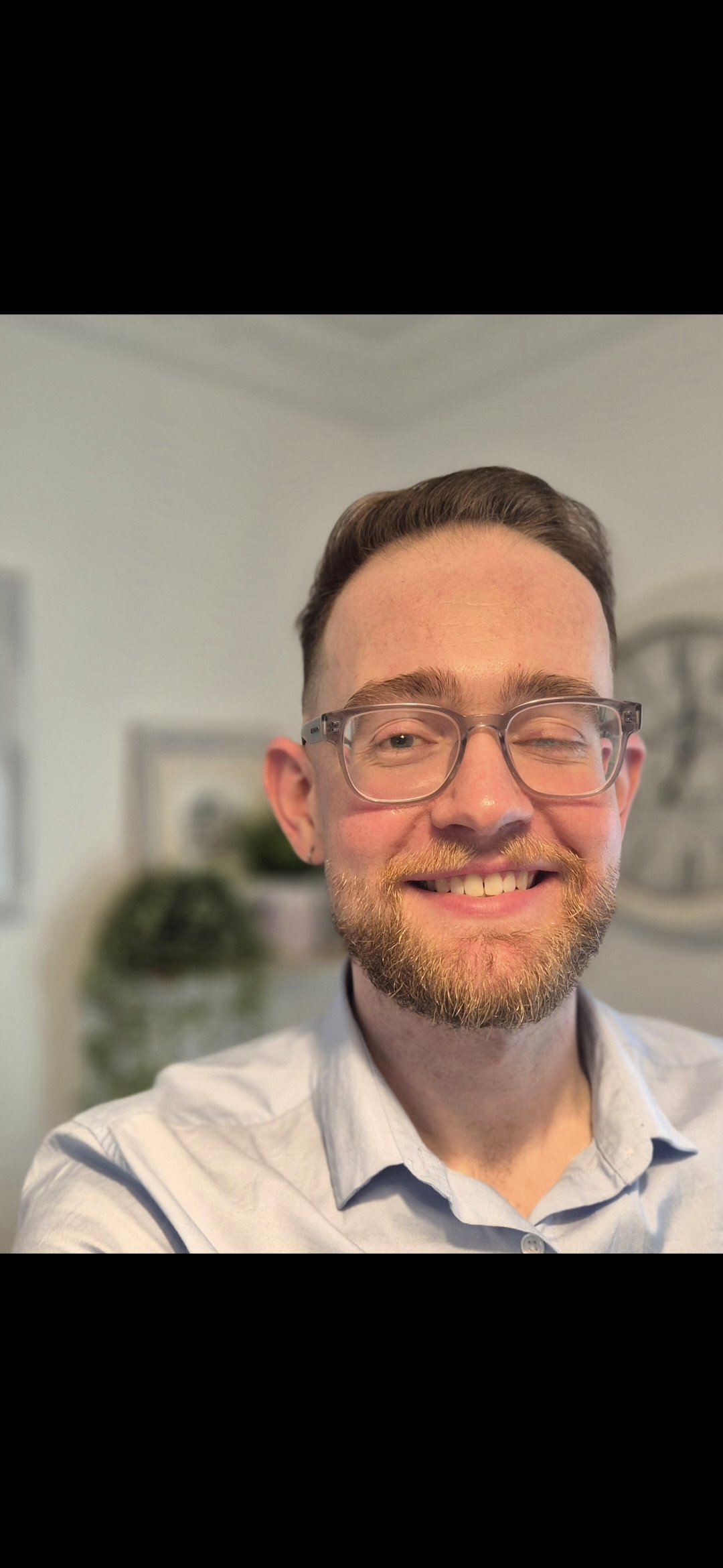 Professional Photograph of Pre-Accredited Psychotherapist Keith Judge in a Light Blue Shirt with a blurred background showing hanging plants and picture frames.
