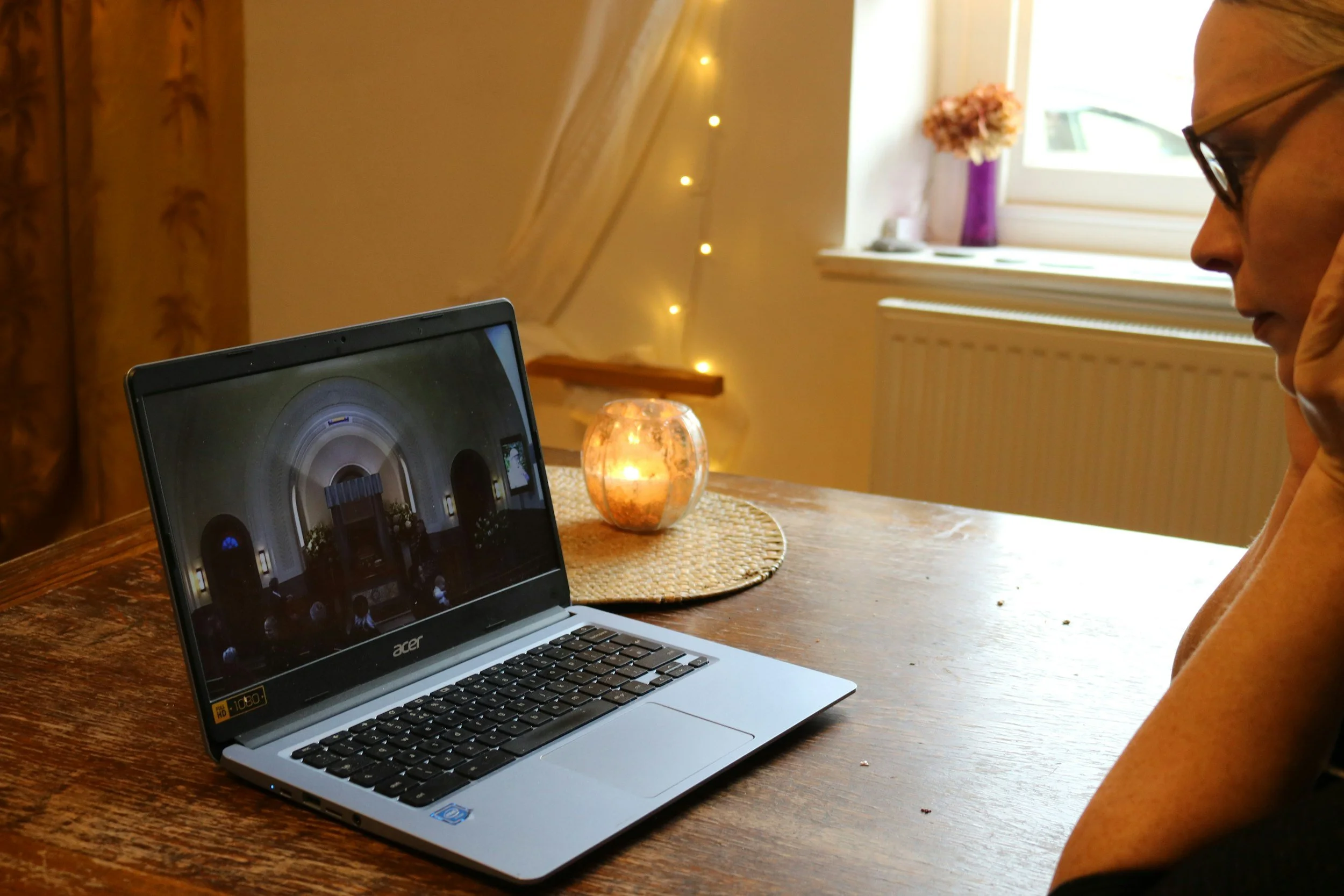 A laptop situated on a beige table with a lit candle placed behind it. There is seemingly hanging LED soft warm lights hanging vertically off the wall near a window ledge.