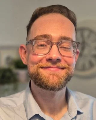 A man with glasses and a beard smiling at the camera in an indoor setting.