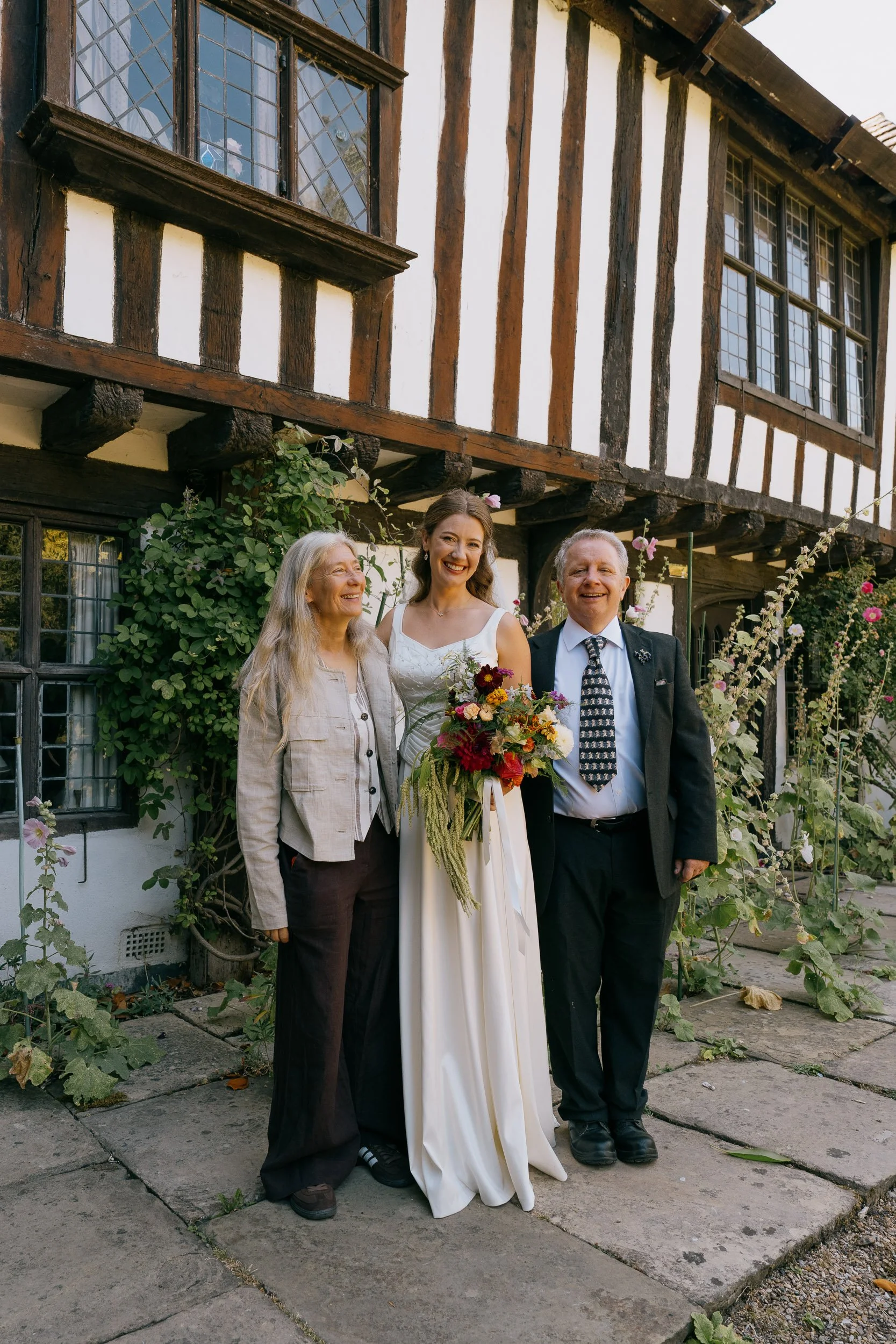 Three people standing outdoors in front of a traditional half-timbered house with white walls and dark wooden beams. The woman in the center is wearing a white wedding dress and holding a bouquet, flanked by an elderly woman and a man, both dressed i