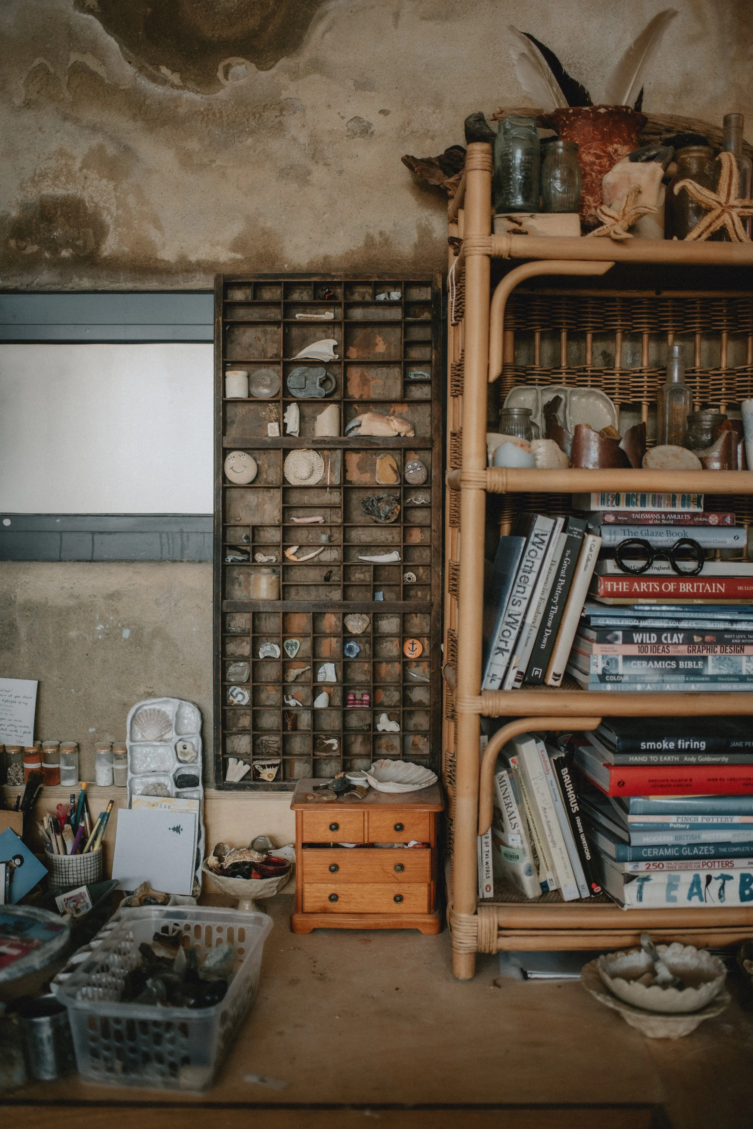 A collection of small jars, seashells, and miscellaneous objects on wooden and wicker shelves, with books and art supplies on a table and shelves in a rustic room with a textured wall.
