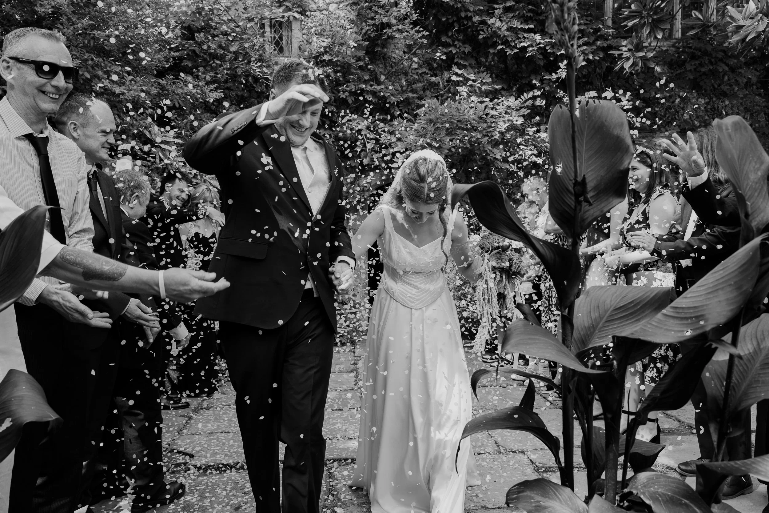Black and white photo of a wedding celebration outdoors with a group of people, including a bride and groom holding hands, surrounded by guests throwing confetti, smiling and celebrating amidst lush greenery.