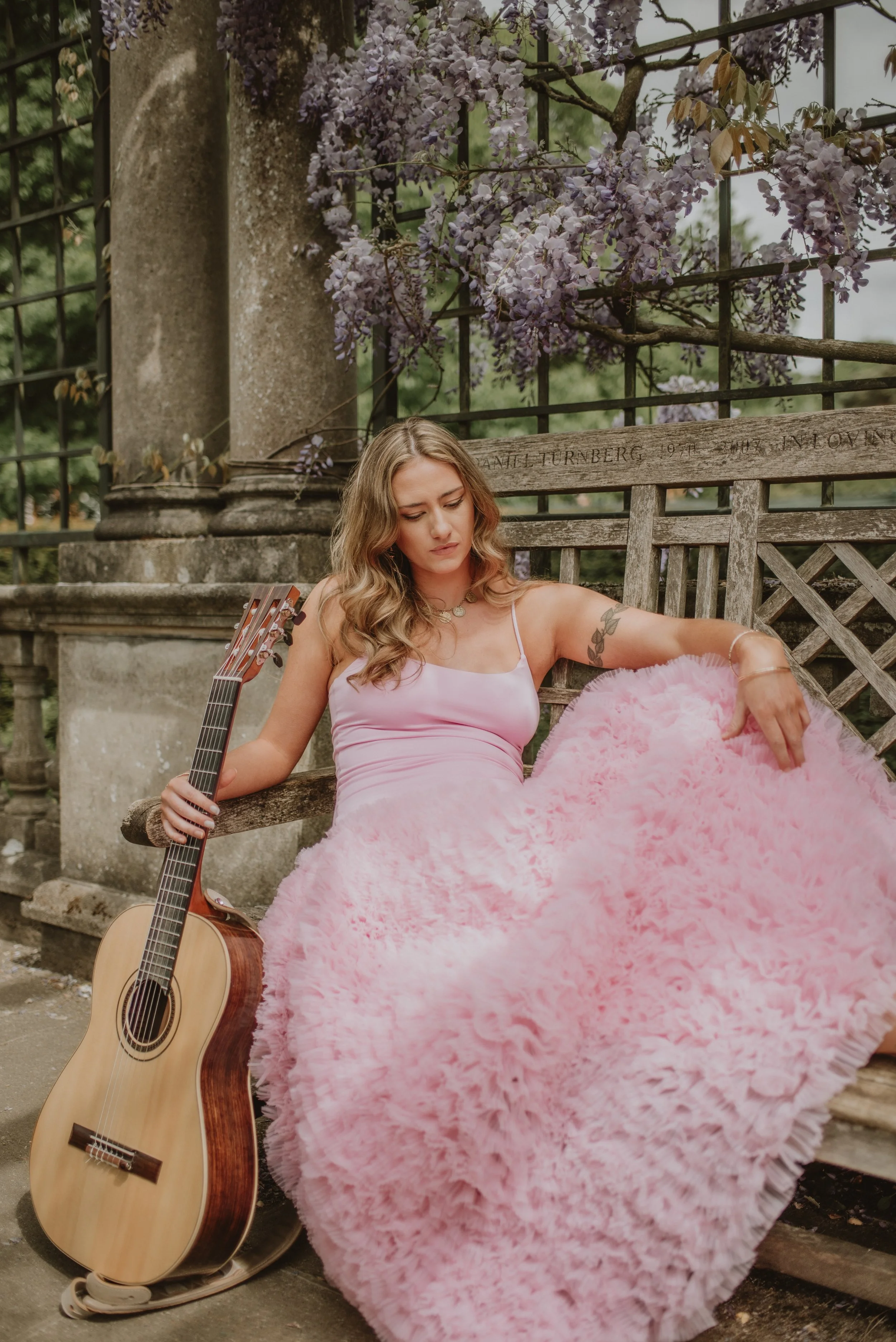 Young woman in a pink dress sitting on a park bench holding a guitar with purple flowering vines in the background.