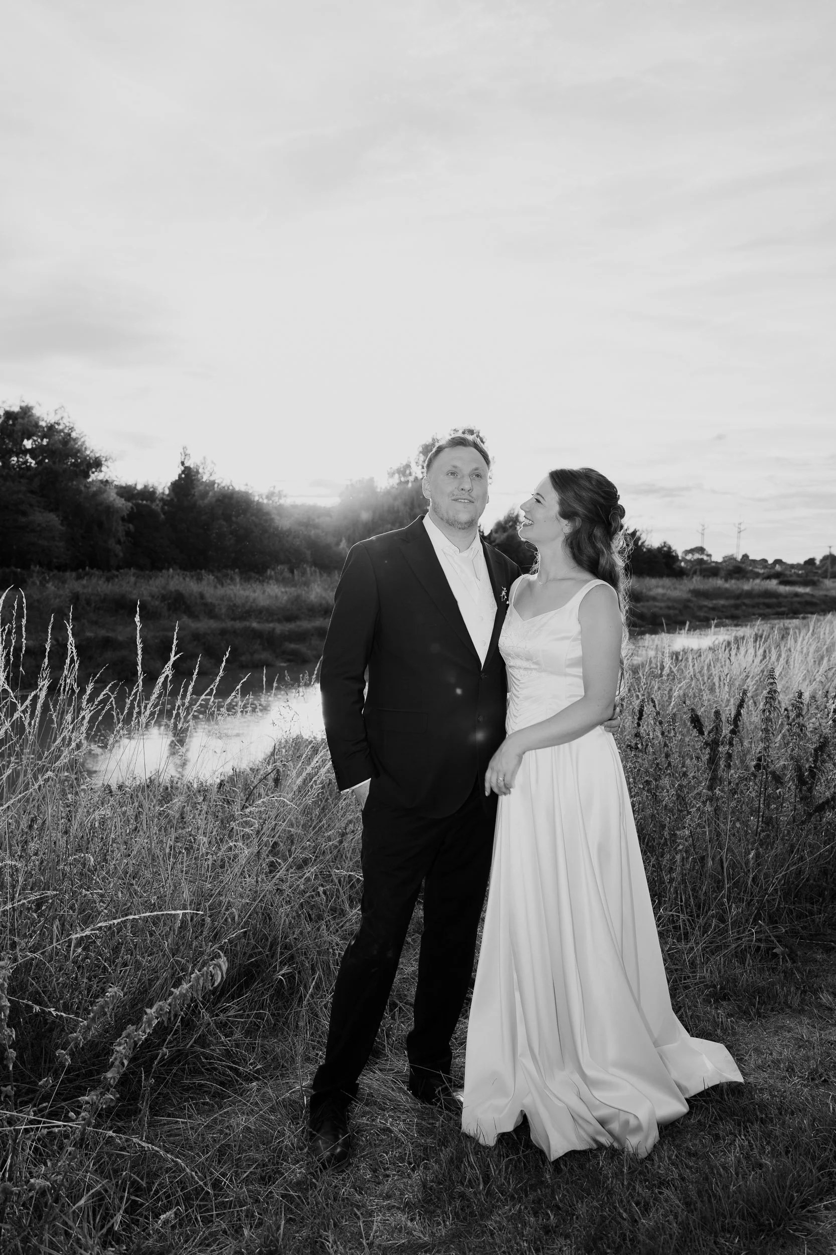 A black and white photograph of a bride and groom standing in a natural outdoor setting, with trees, grass, and a small water body behind them, during sunset.