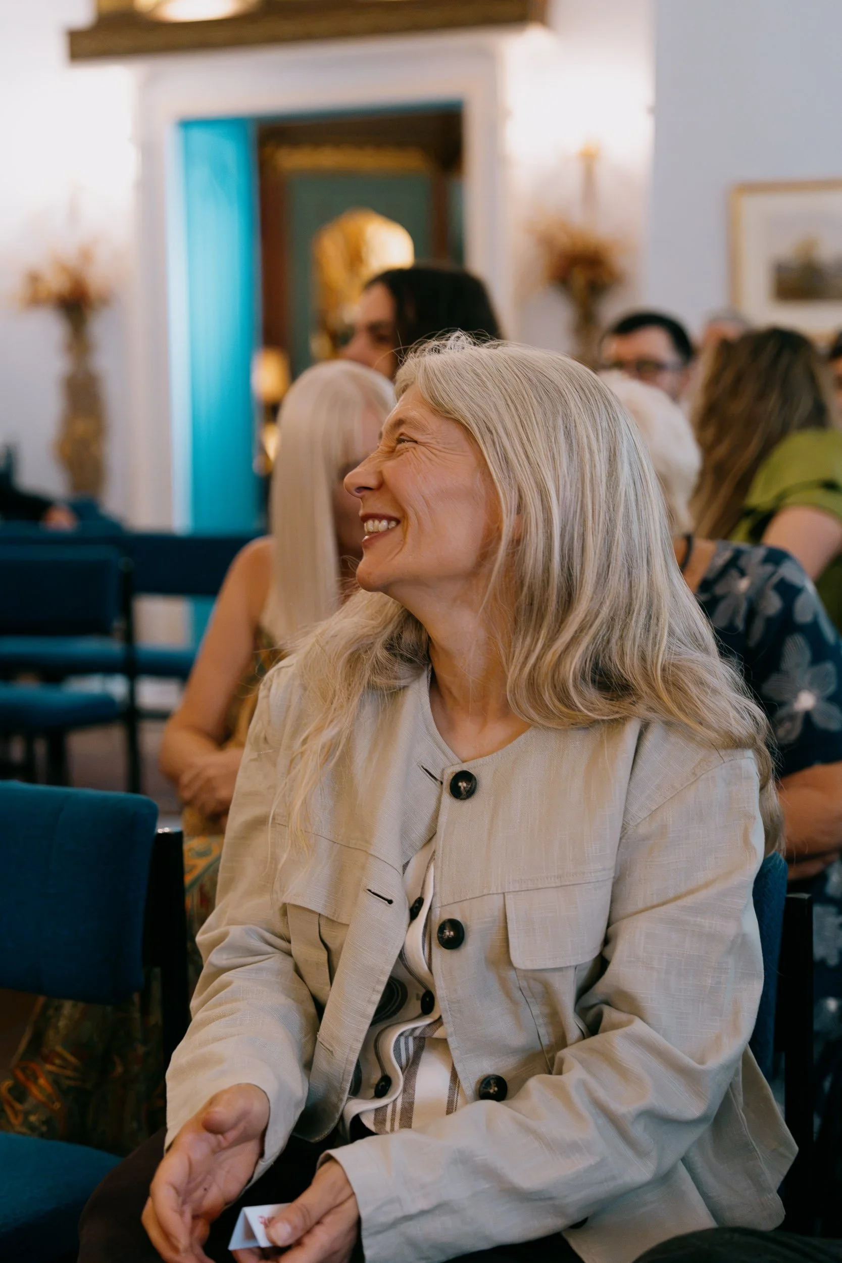 An elderly woman with long gray hair smiling while sitting at a social gathering in a decorated room, wearing a beige jacket and holding a small paper object.