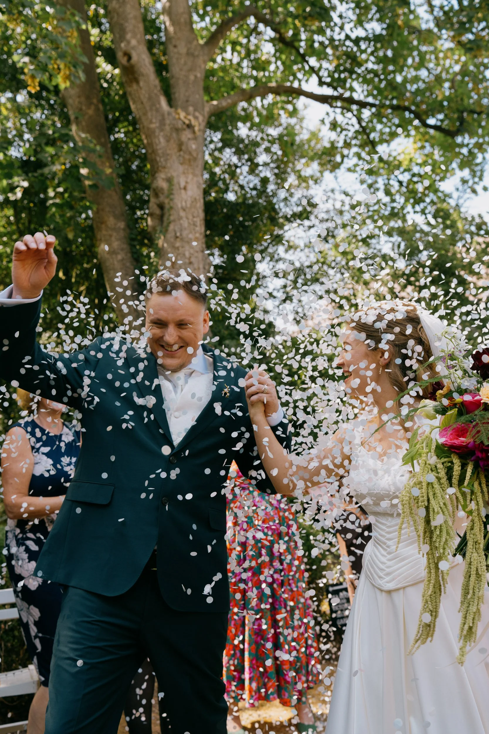 A man in a tuxedo and a woman in a wedding dress celebrating outdoors, surrounded by falling confetti, with trees and greenery in the background.