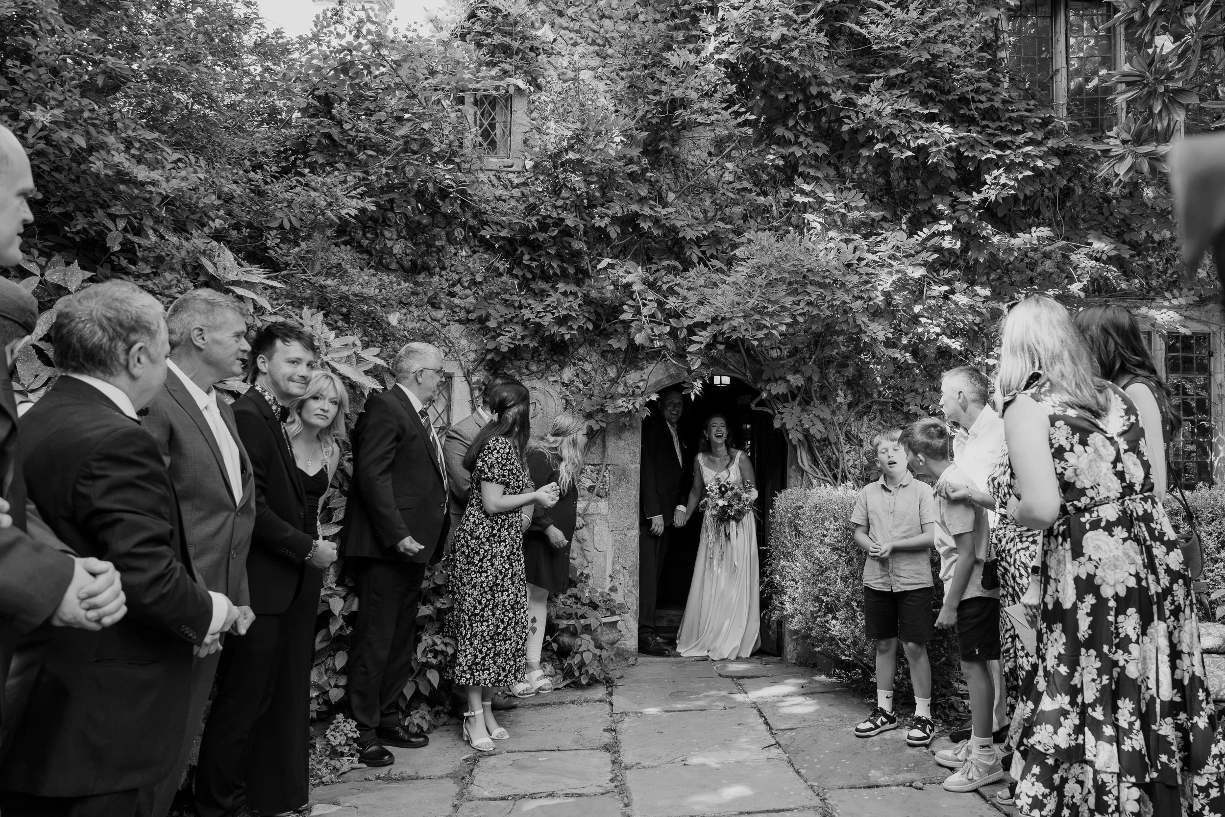 People attending a wedding ceremony outside in front of a building covered with ivy and foliage, with some guests standing and observing.