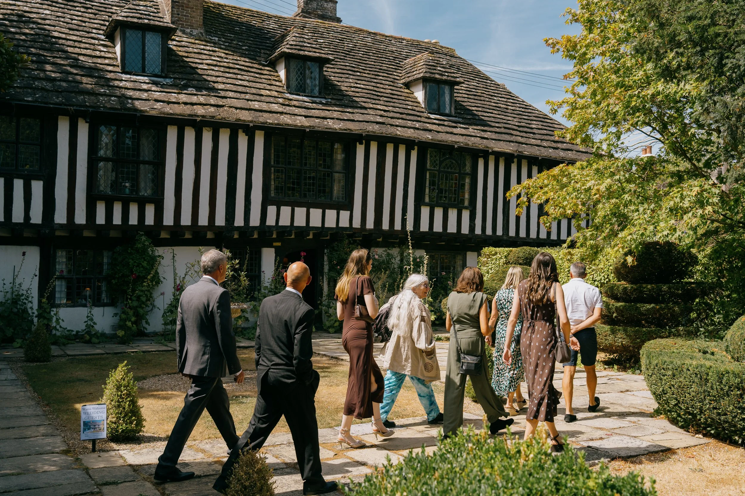 A group of wedding guests walking outside a large, historic, black-and-white timber-framed house surrounded by garden and trees on a sunny day.