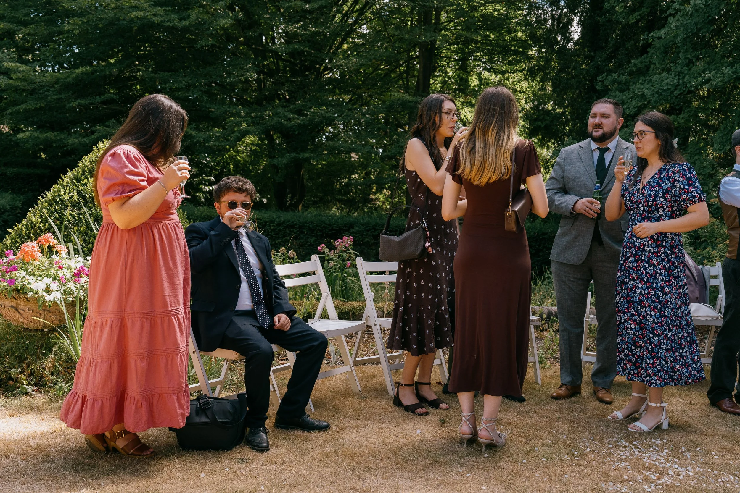A group of seven people, six women and one man, gathered outdoors in a garden during daytime. Some are standing while one woman is seated. They are dressed in semi-formal attire, holding glasses of wine or champagne, and appear to be socializing, wit