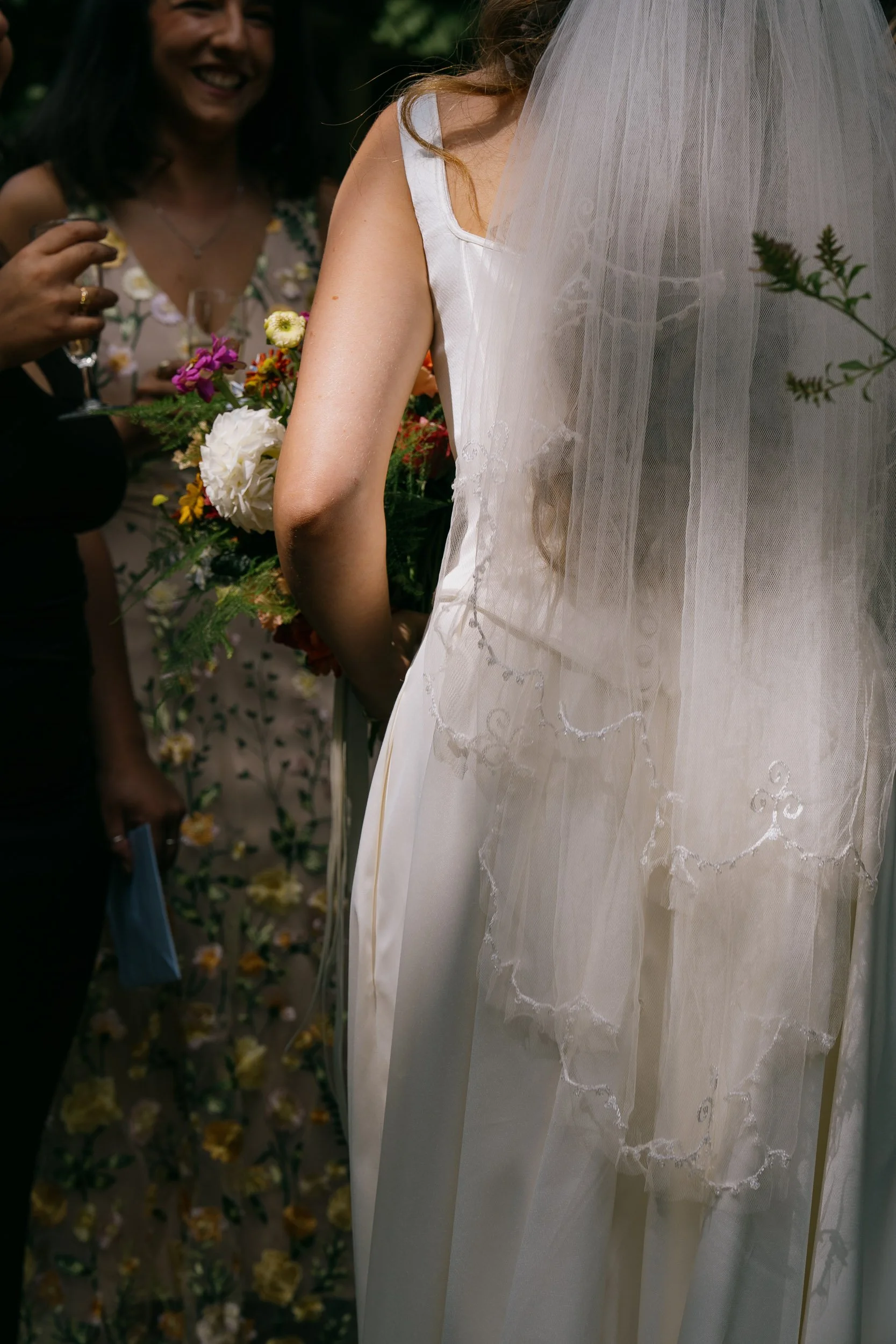 A bride in a white dress with lace detail is holding a bouquet of colorful flowers. She is wearing a veil with embroidered swirling patterns. A woman in a floral dress is smiling and holding a glass, standing close to the bride. The background is blu