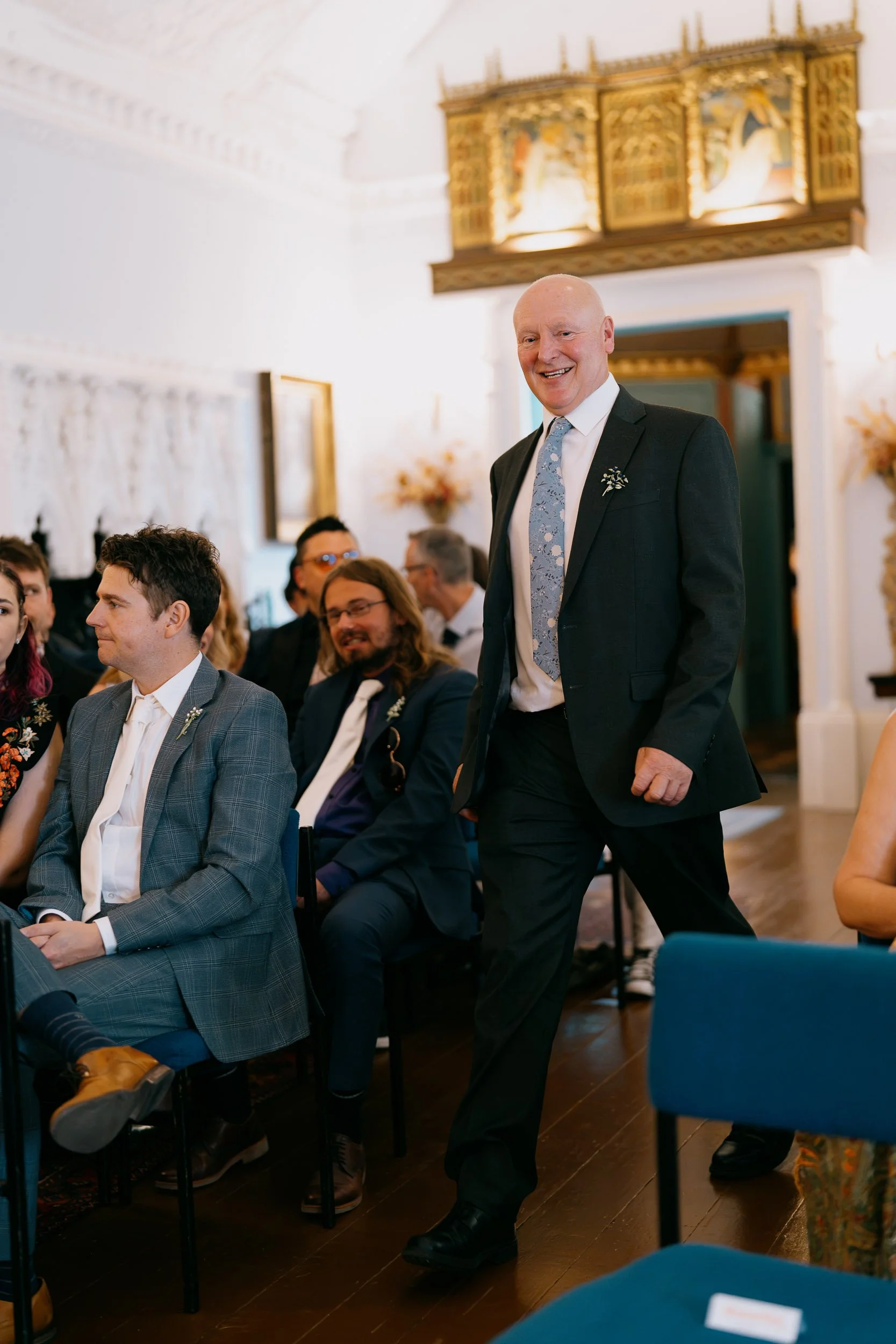 A man in a dark suit with a gray tie walking through a room with several seated guests. The room has white walls, ornate decorations, and golden artwork on the walls.
