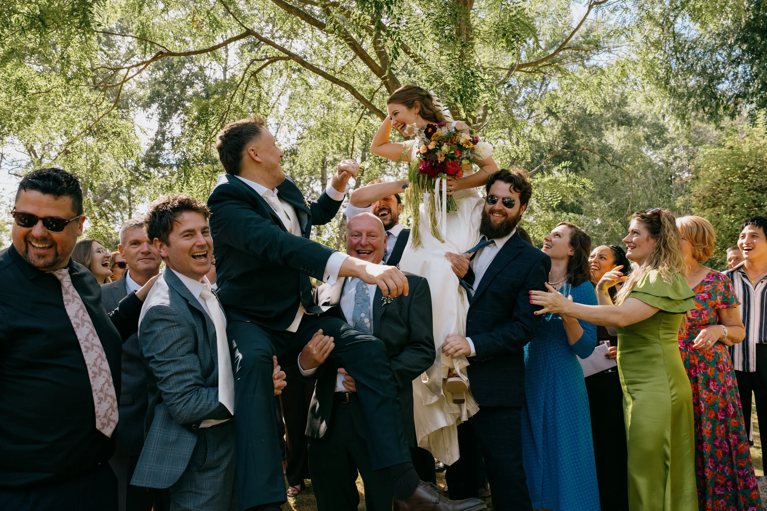 A group of wedding guests outdoors, with a bride sitting on a man's shoulders, surrounded by laughing people in colorful attire.