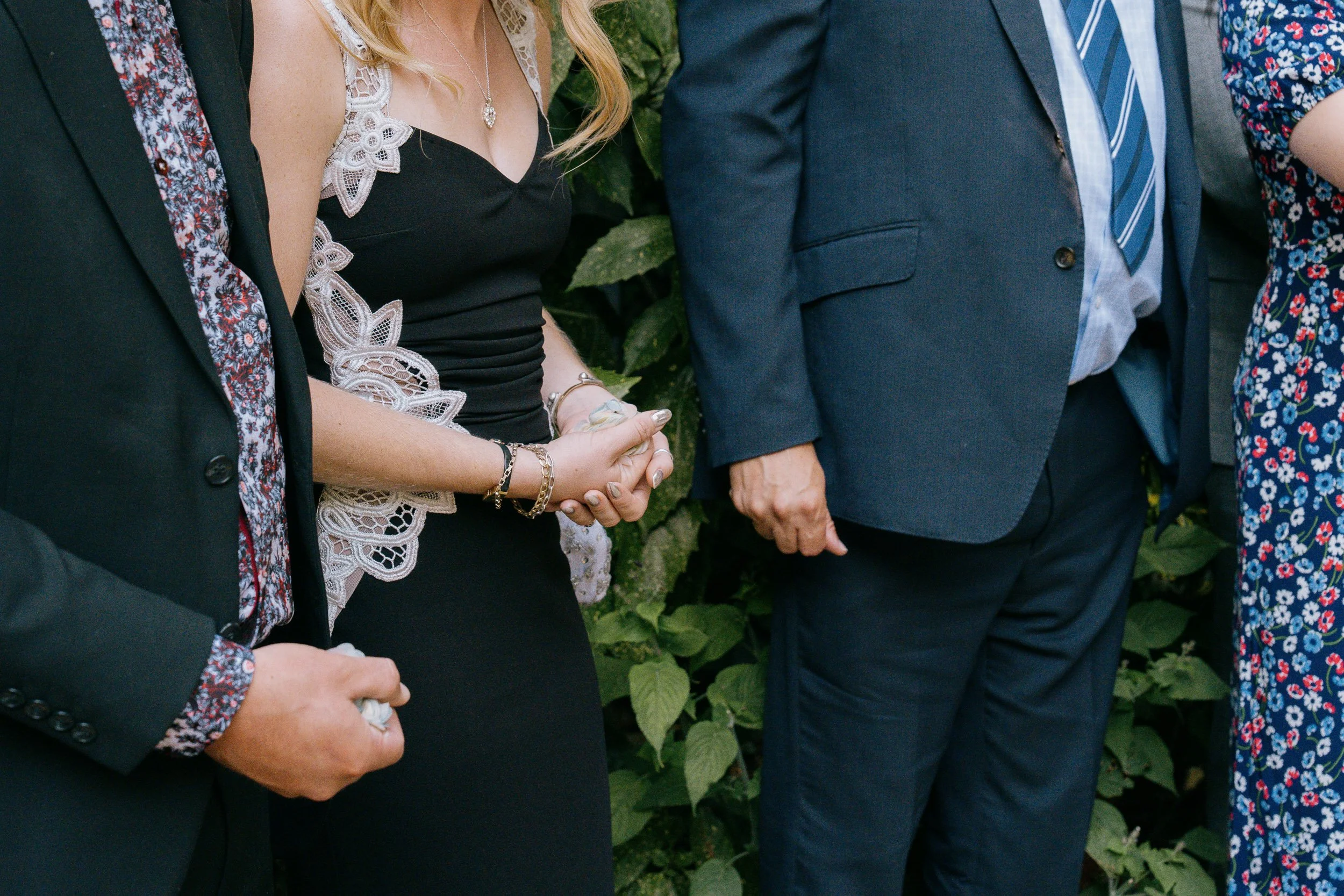 People dressed in formal attire standing close together outdoors, holding hands.