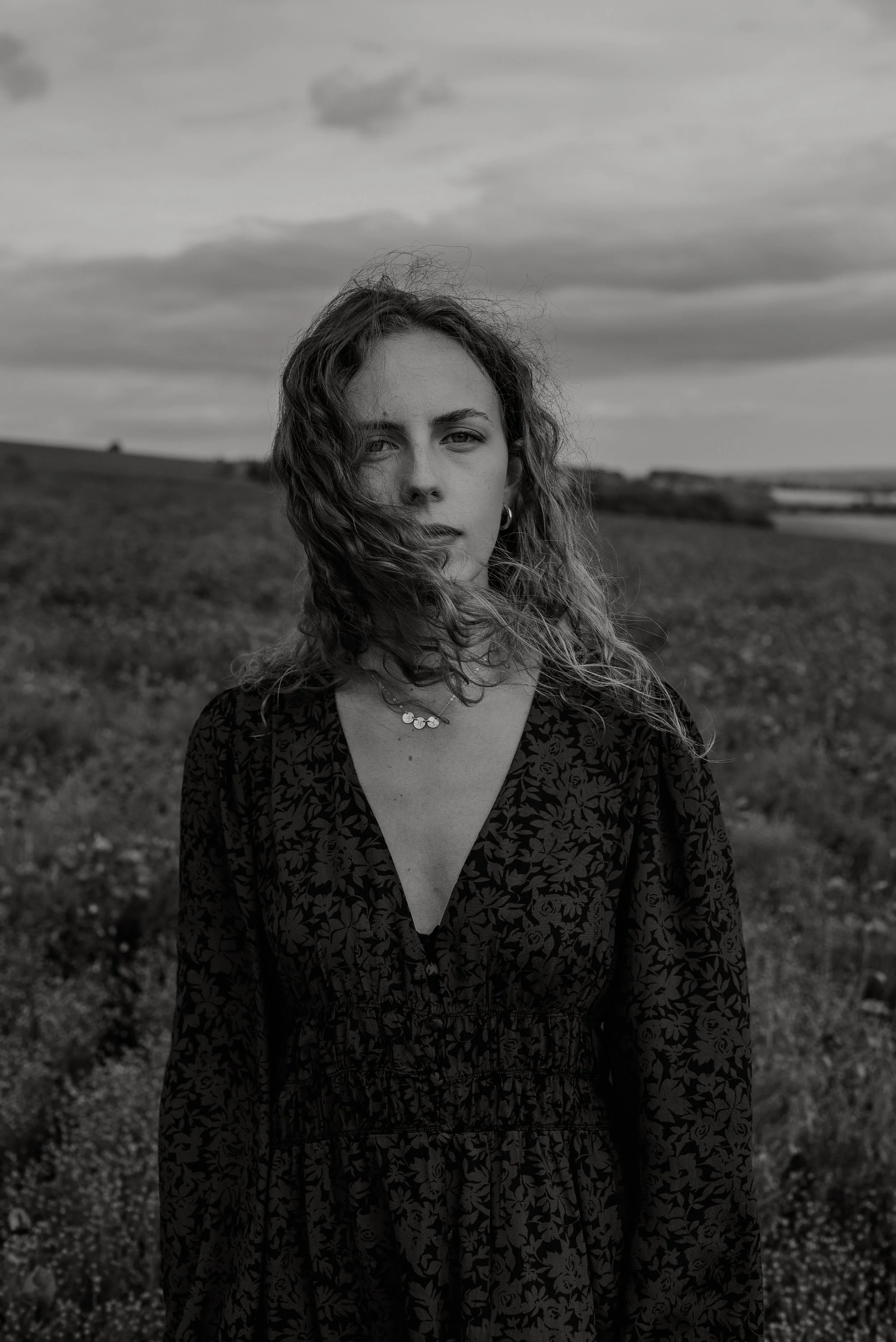 A woman with wavy hair standing outdoors in a field with a cloudy sky in the background, wearing a patterned dress and a necklace.