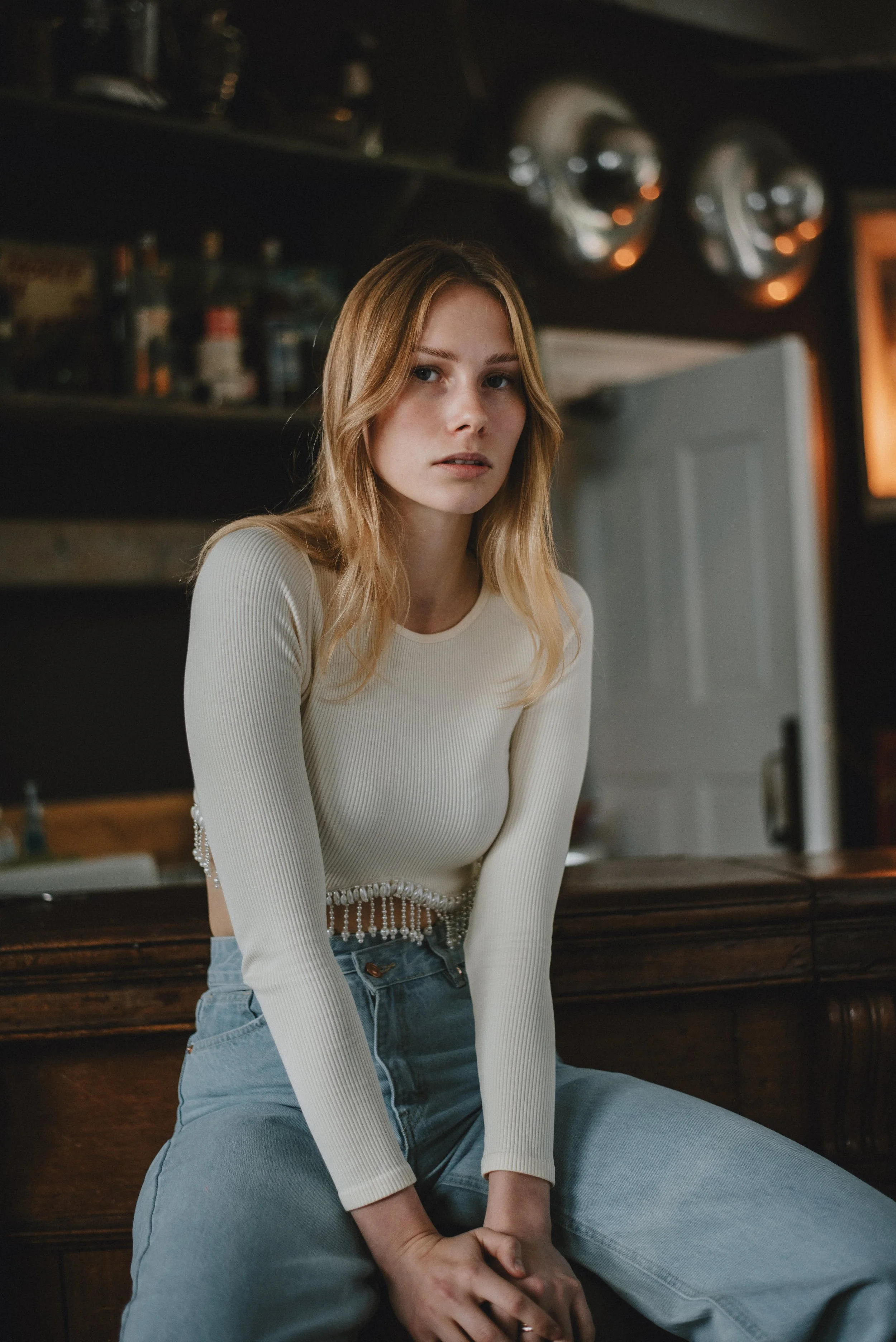 A young woman with blonde hair sits on a wooden bar counter in a dimly lit room, wearing a beige long-sleeve top and blue jeans.