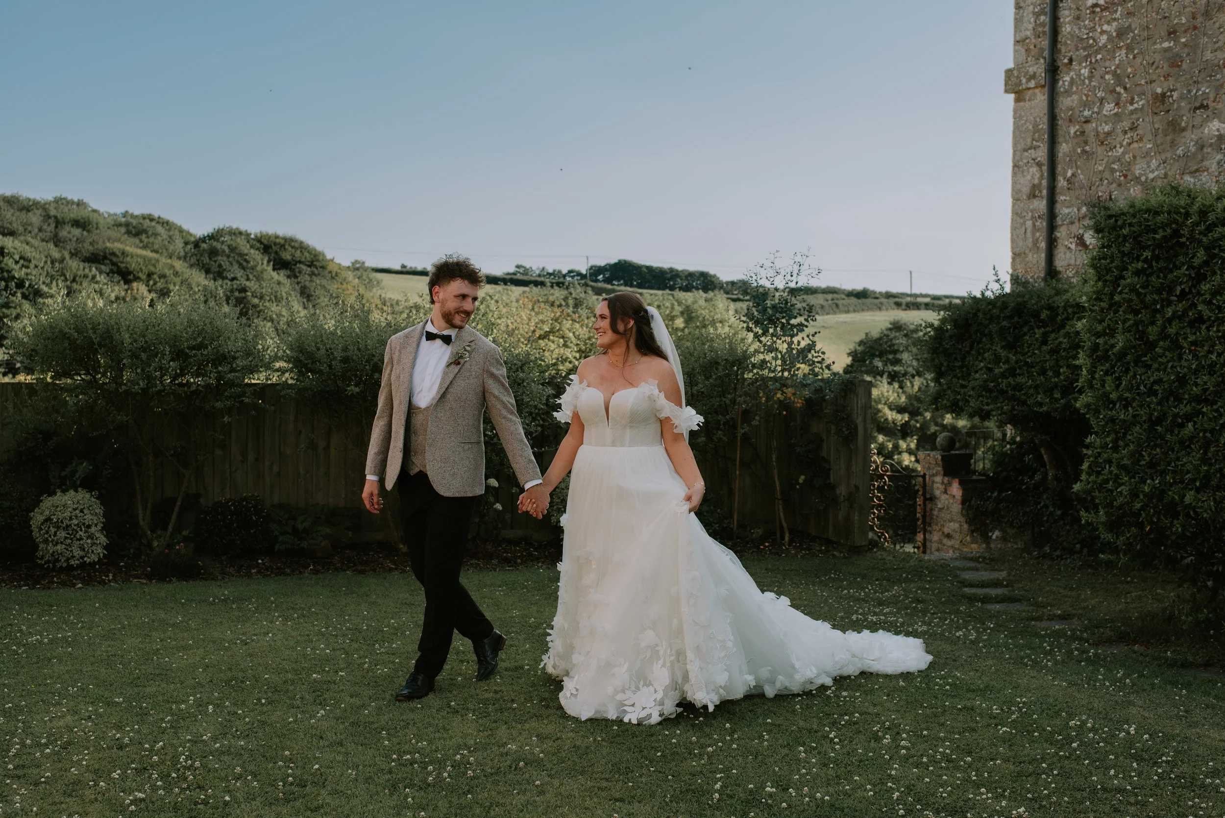A bride and groom walking hand in hand on a grassy yard, smiling at each other, with greenery and a stone building in the background on a sunny day.
