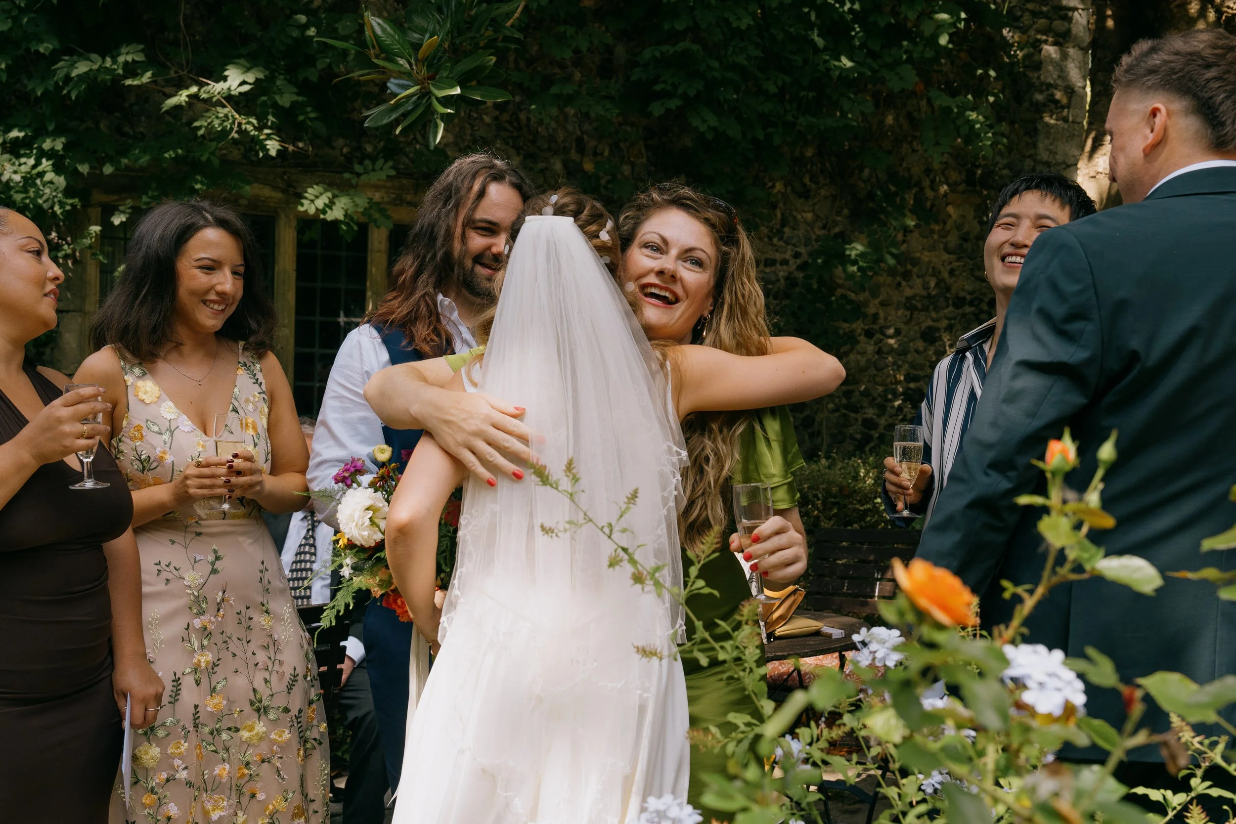 A bride in a white wedding dress hugging a woman. Around them, several women and a man in a suit are smiling and holding drinks outdoors, with greenery and flowers in the background.
