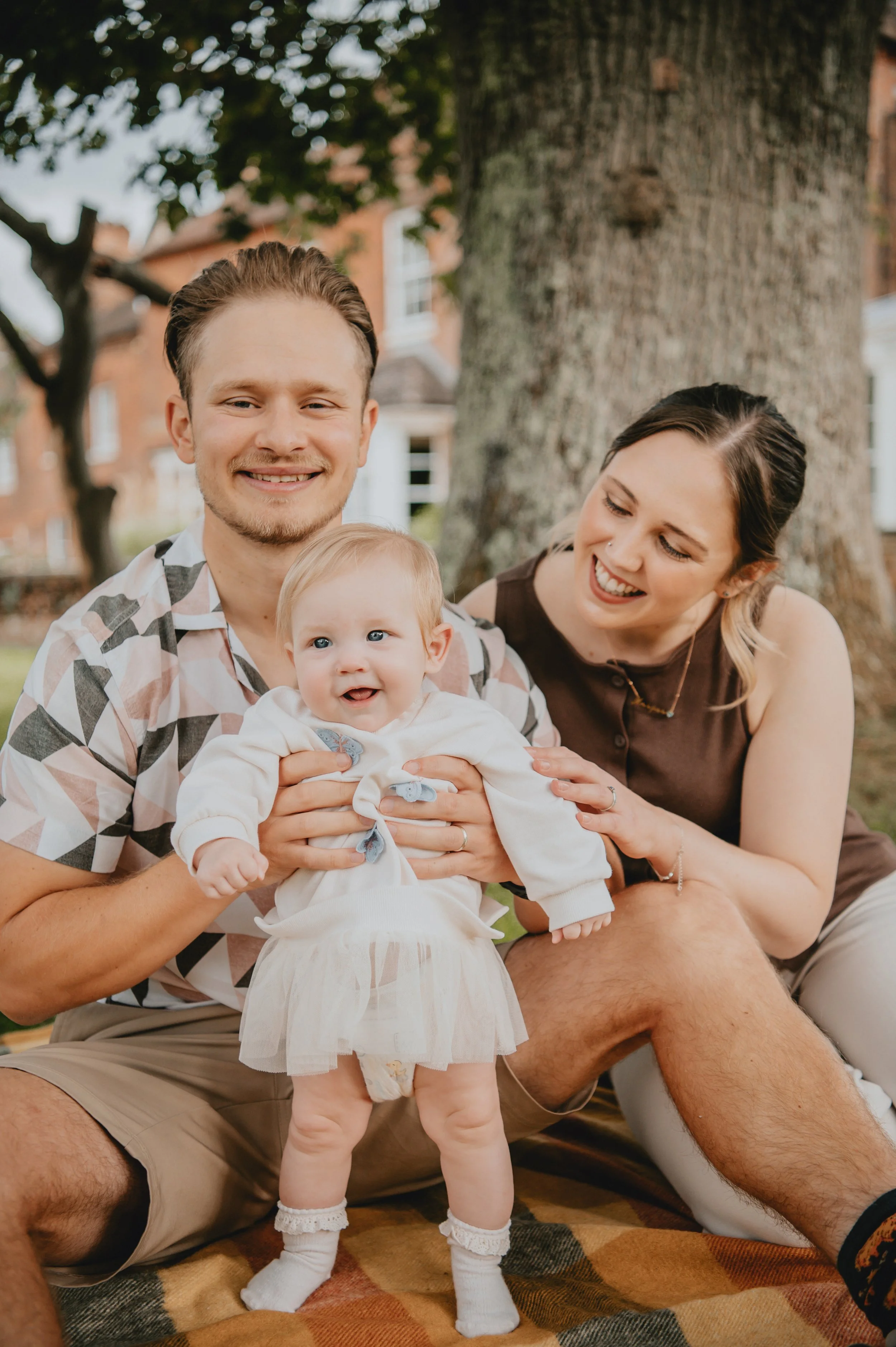 A smiling couple with a baby girl outdoors near a large tree, with houses in the background, sitting on a blanket.