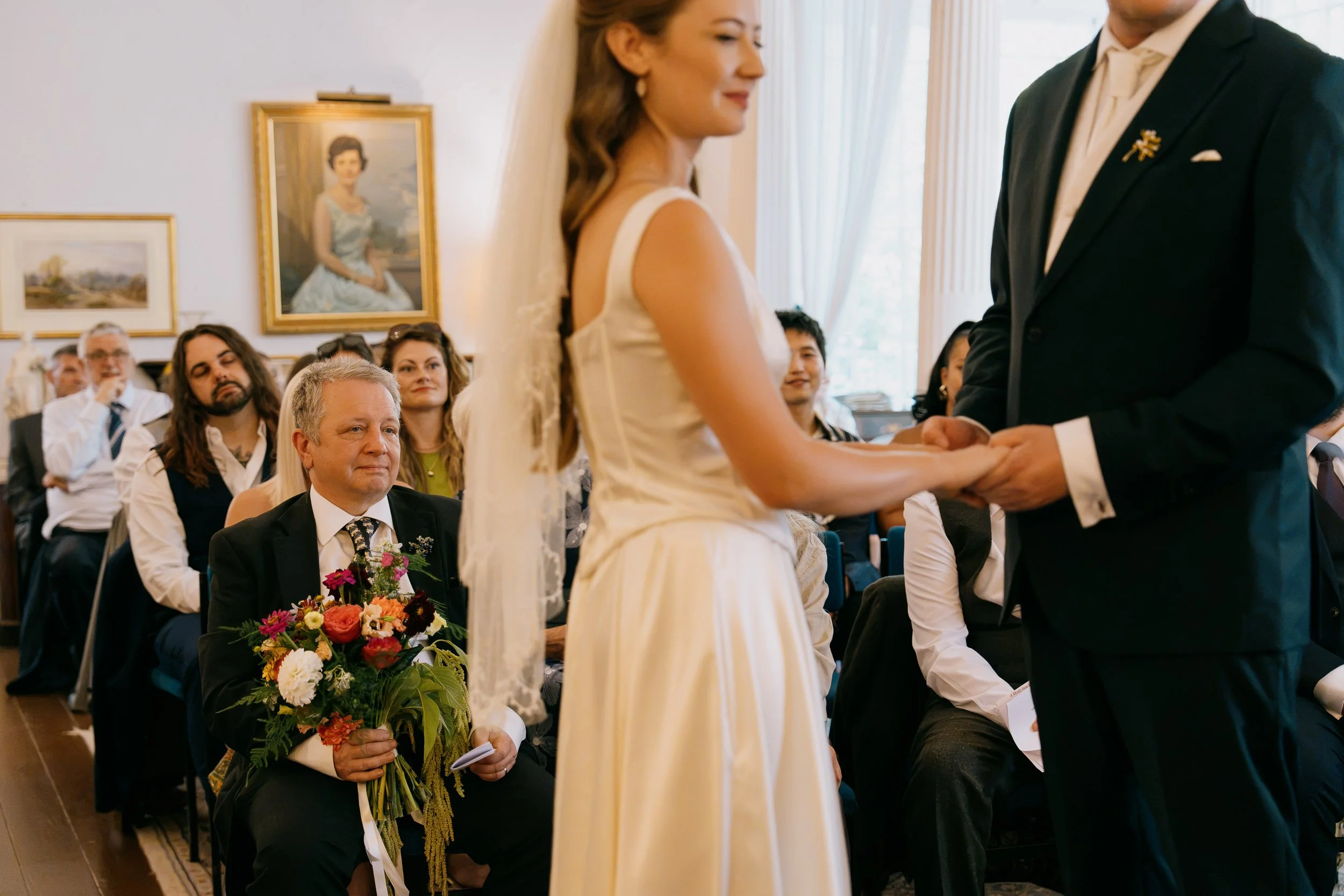 A bride and groom holding hands during a wedding ceremony with seated guests watching, some with flowers and formal attire, in a room with artwork on the wall.