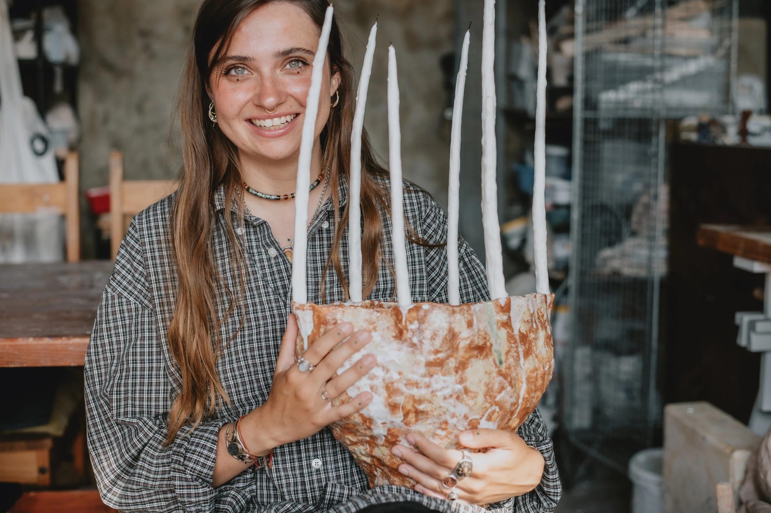 A young woman with long brown hair, earrings, multiple rings, and a necklace, holding a large rock with white sticks attached to it, in an indoor workshop or studio setting.