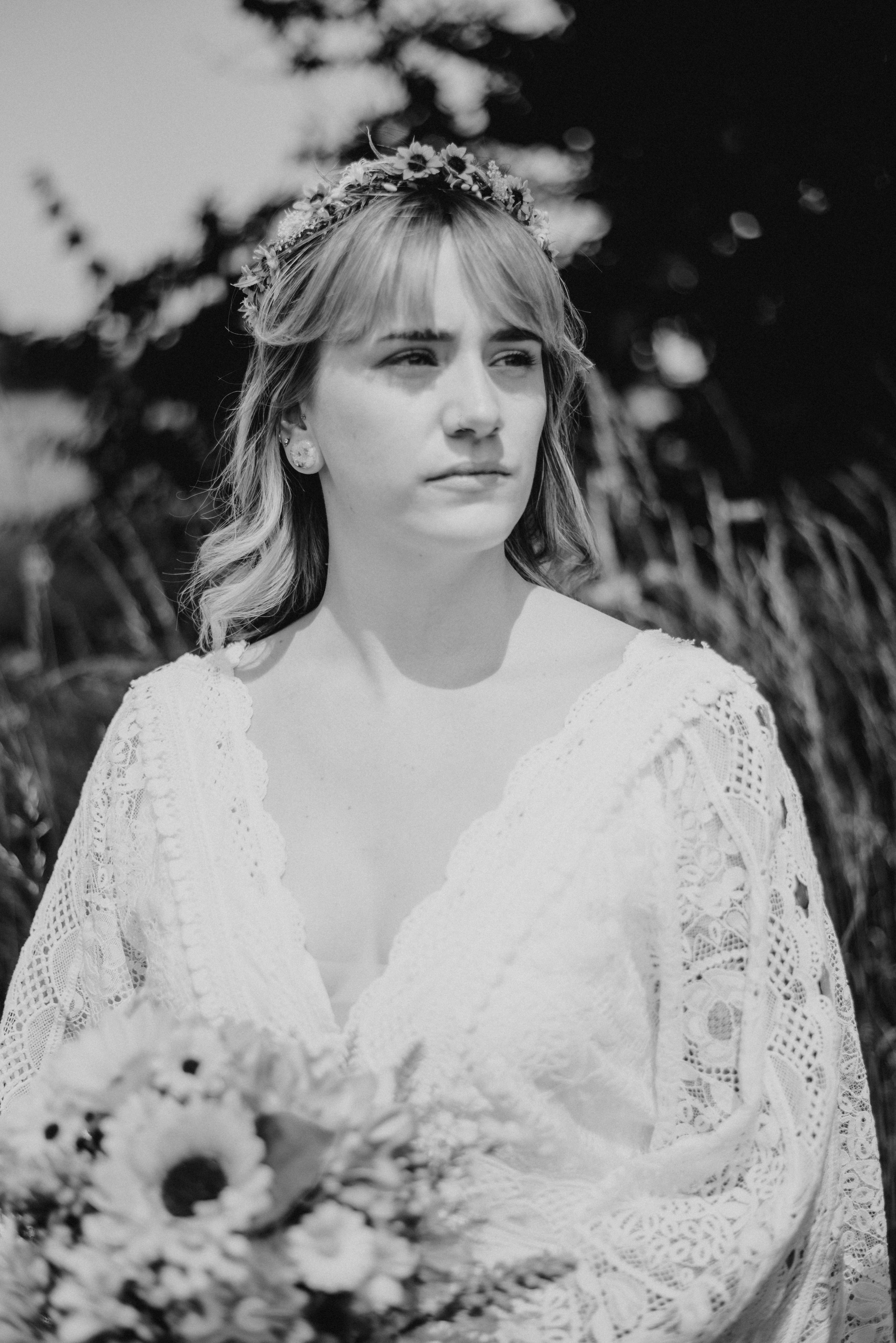 Black and white photo of a young woman with a floral crown, holding a bouquet of flowers, outdoors with trees in the background.