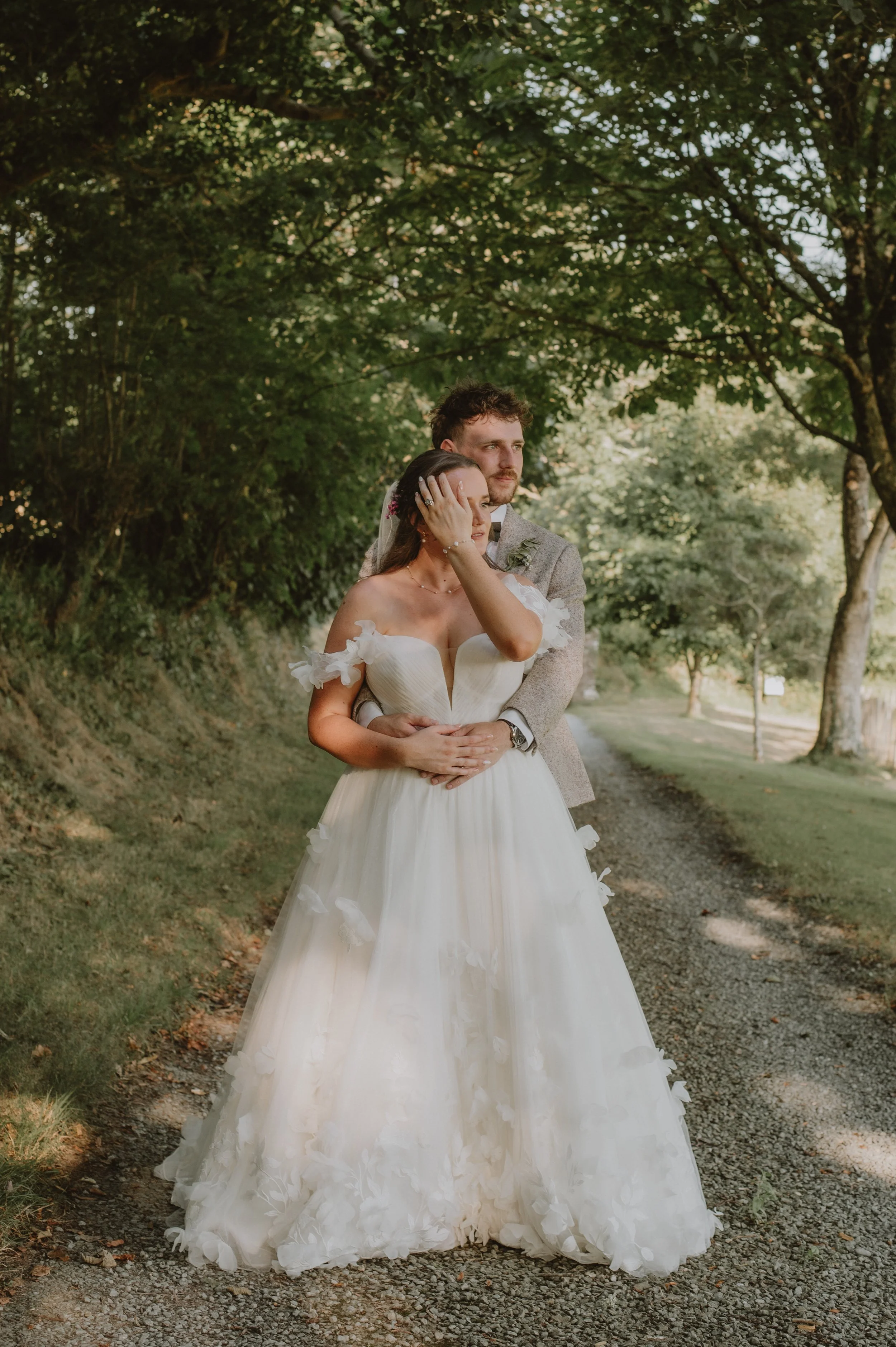 A bride and groom hugging outdoors on a shaded dirt path among green trees during their wedding.