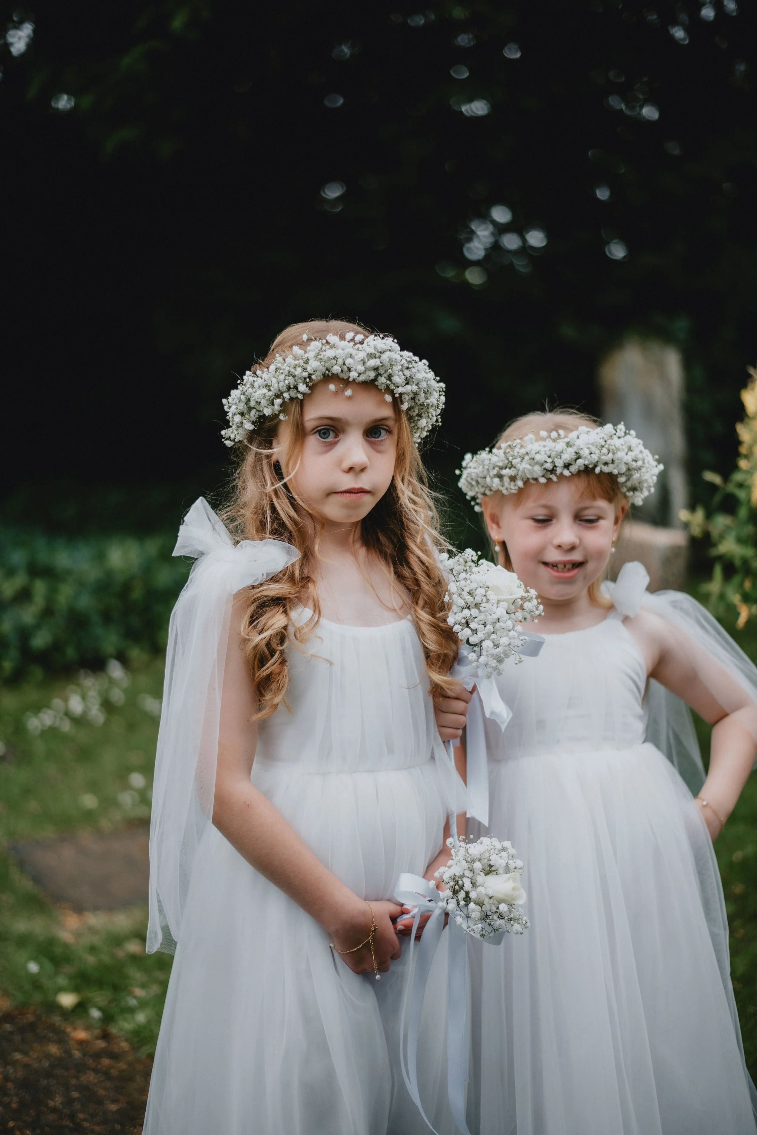 Two young girls dressed in white dresses with flower crowns and holding small bouquets of white flowers, standing outdoors in a garden.