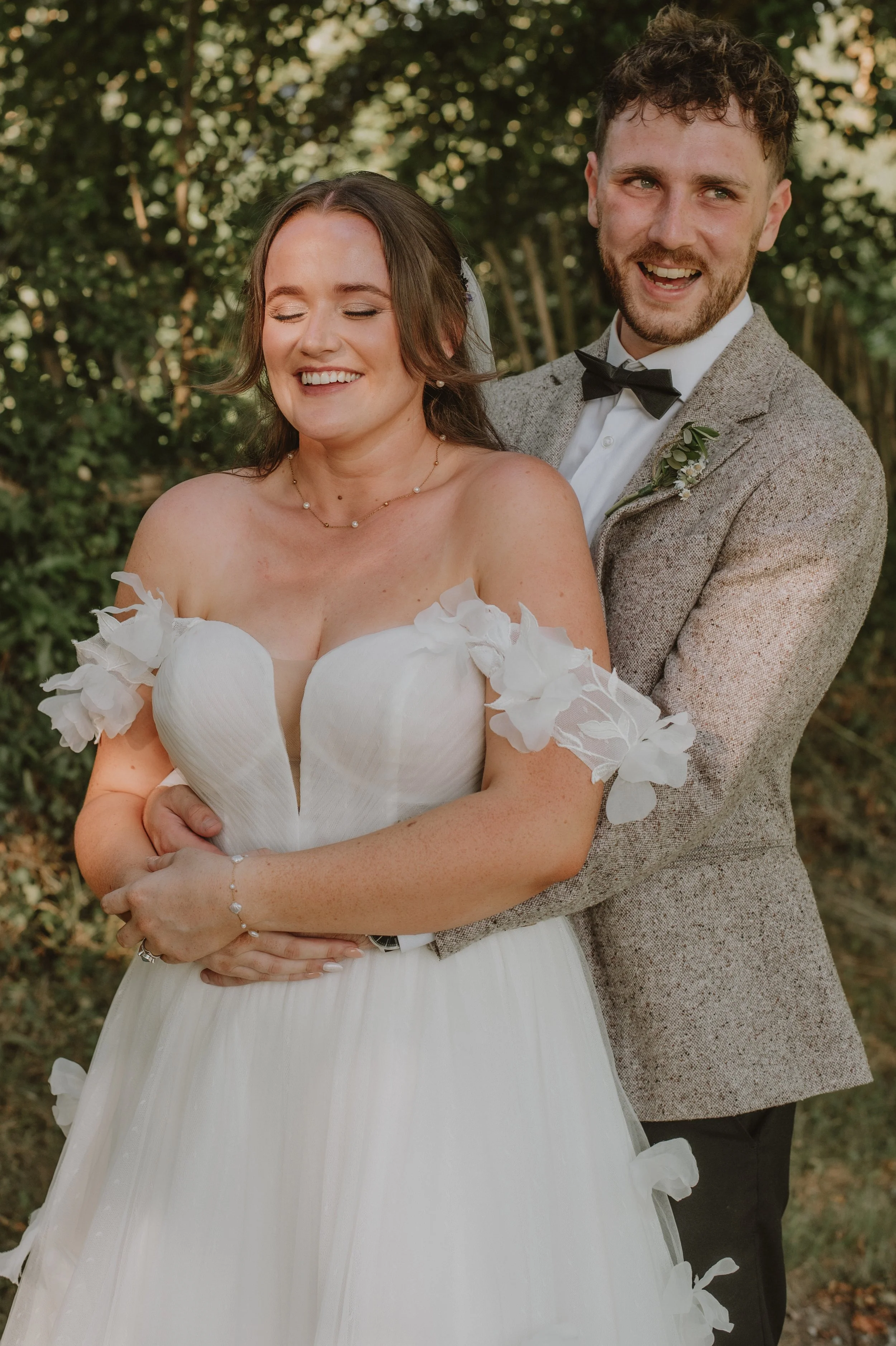 A smiling bride and groom stand outdoors, with the bride wearing a white wedding dress with floral accents on the shoulders, and the groom in a tan suit with a bow tie, embracing each other.