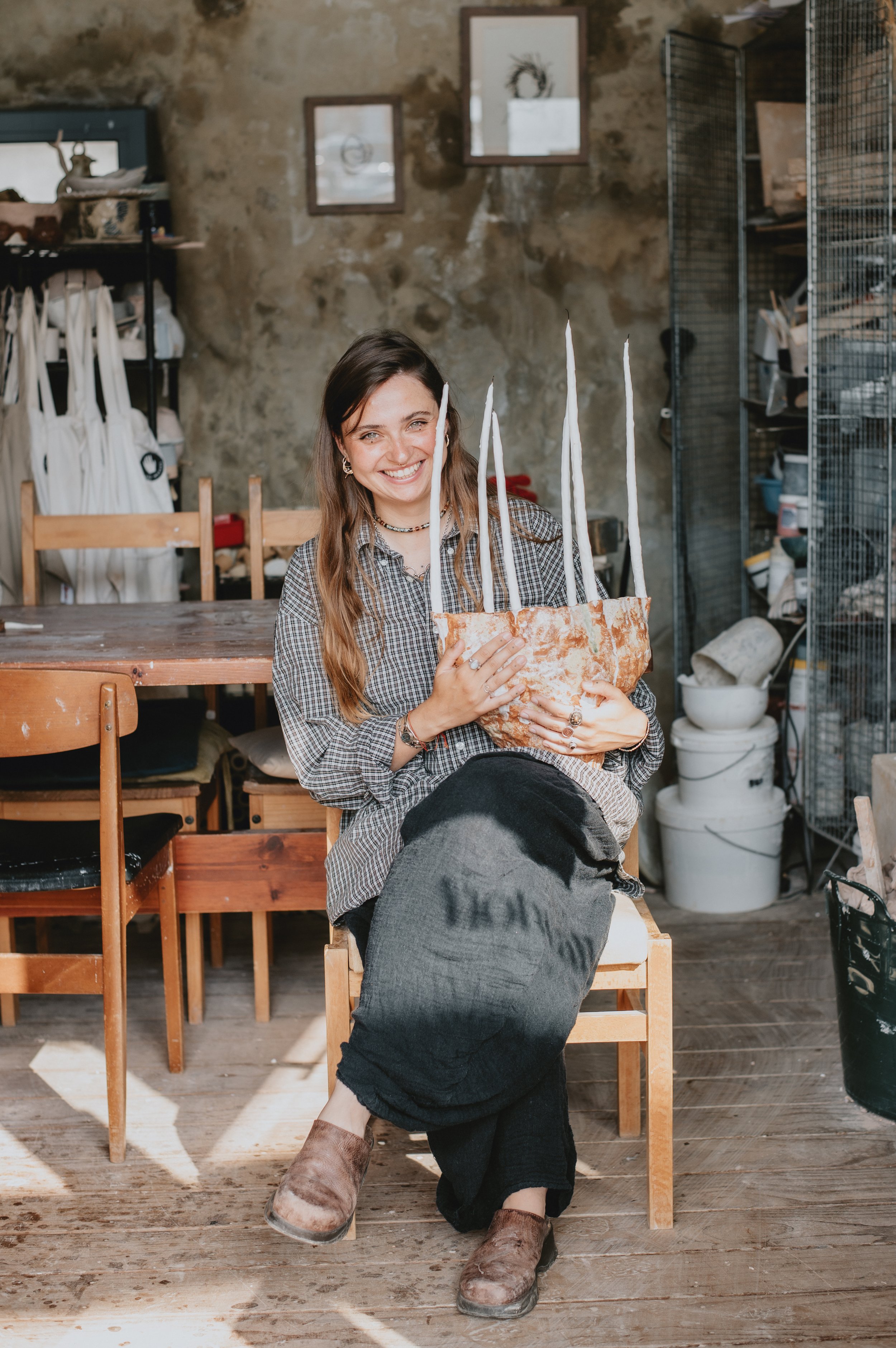 Woman smiling and holding a large, rustic brown paper bag with tall white candles inside in a cozy workshop or studio.