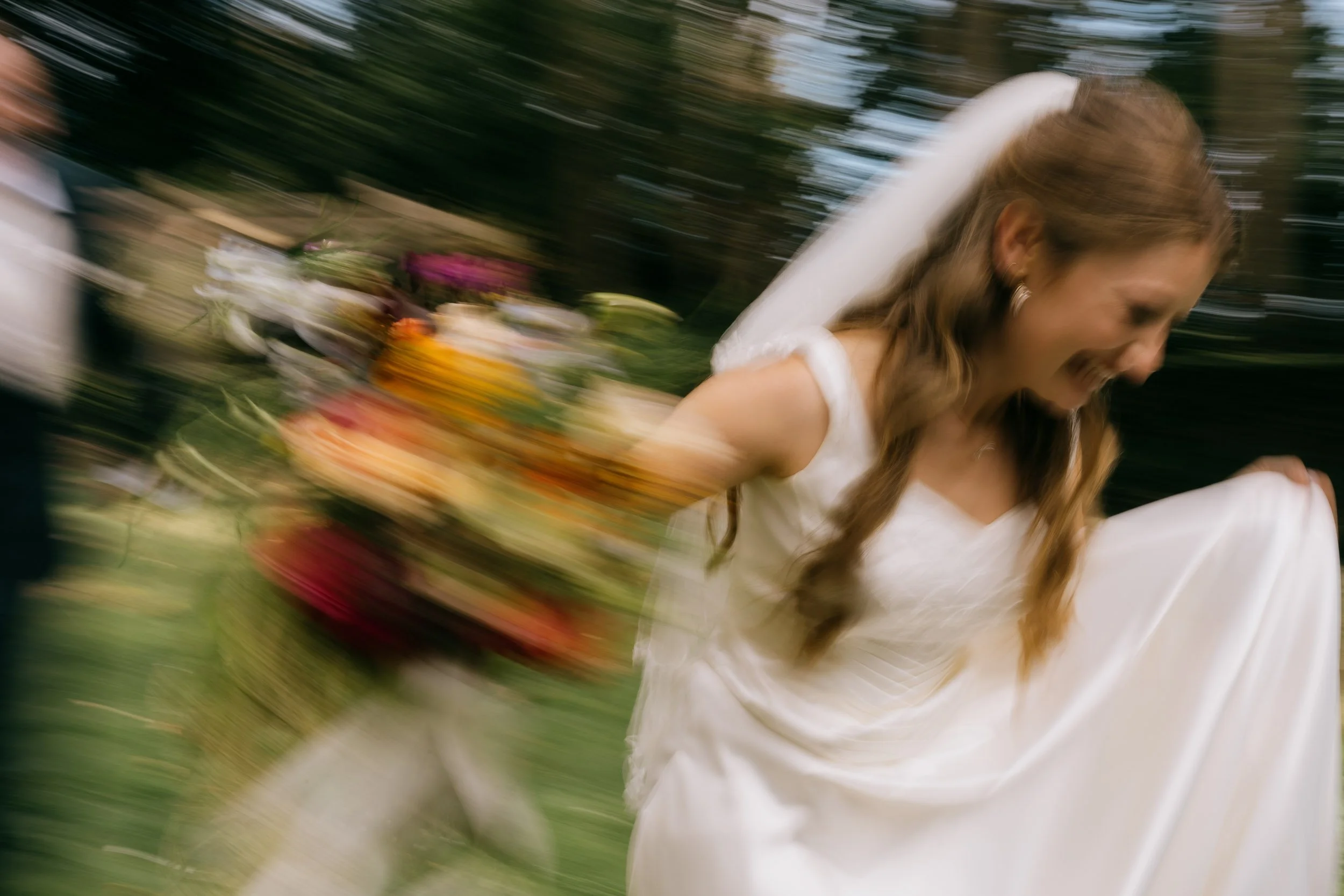 A woman with long hair wearing a lace white dress and a veil, smiling as she walks outdoors with a bouquet of flowers behind her, with motion blur indicating movement.