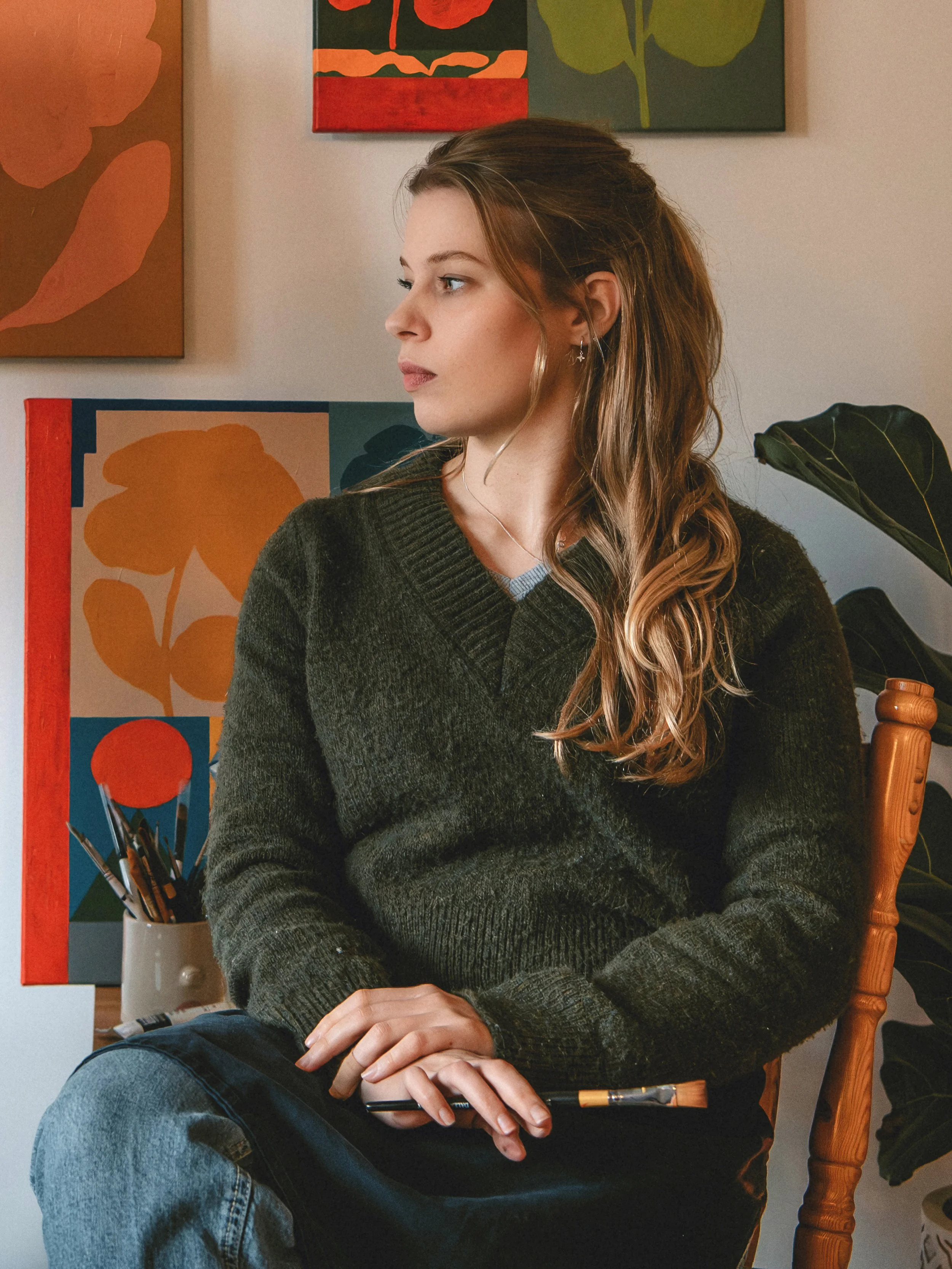 A woman with long wavy hair sitting in a wooden chair, looking to her left, in front of colorful abstract paintings and a large green plant.