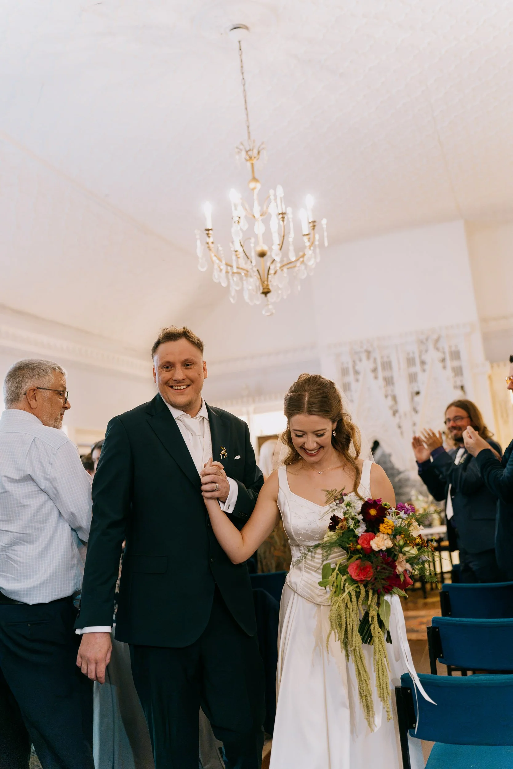 A newlywed couple dancing and smiling at their wedding reception, surrounded by guests in a decorated room with a chandelier on the ceiling.