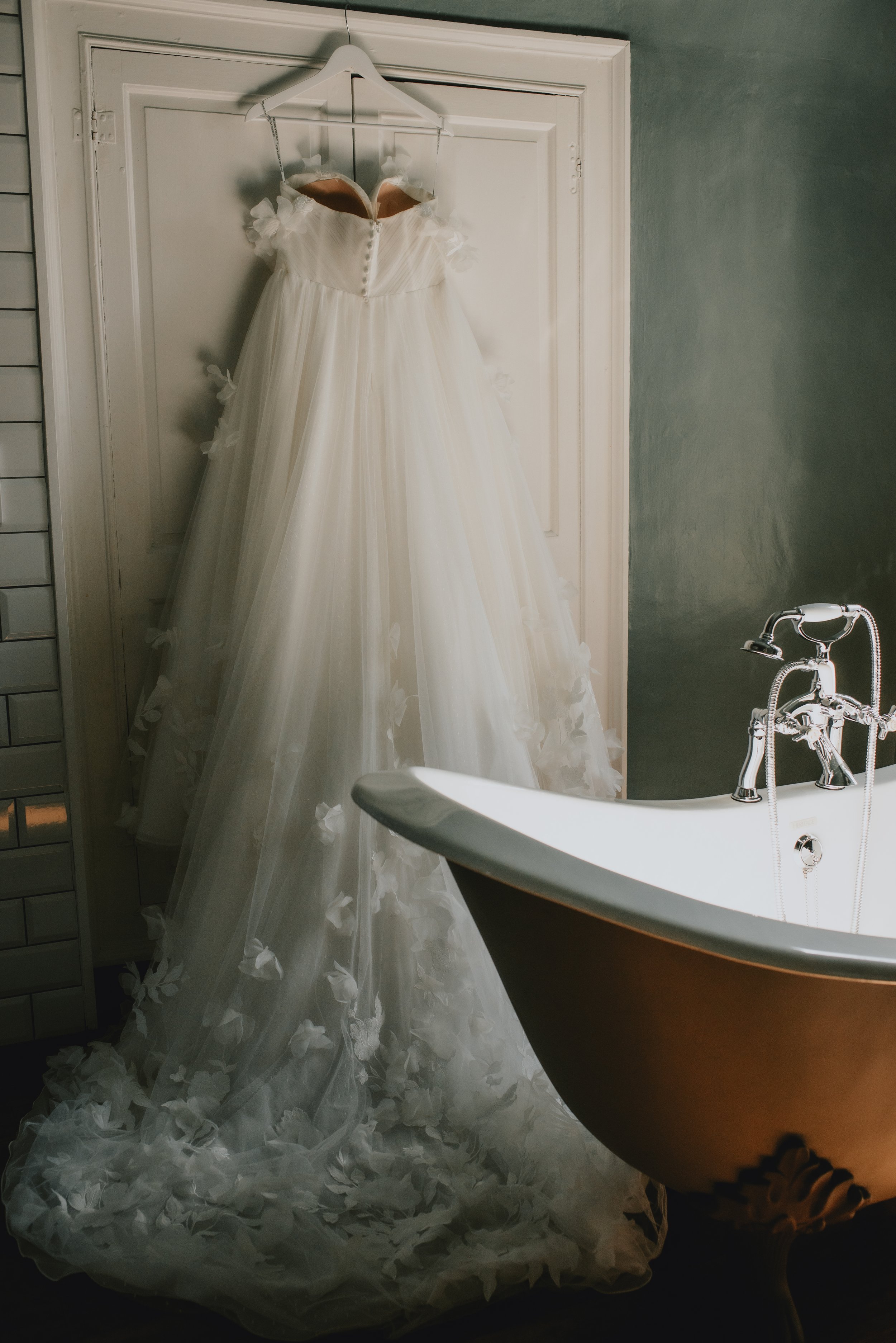 A white wedding dress hanging on a hanger on a door, with a vintage bathtub nearby in a bathroom.