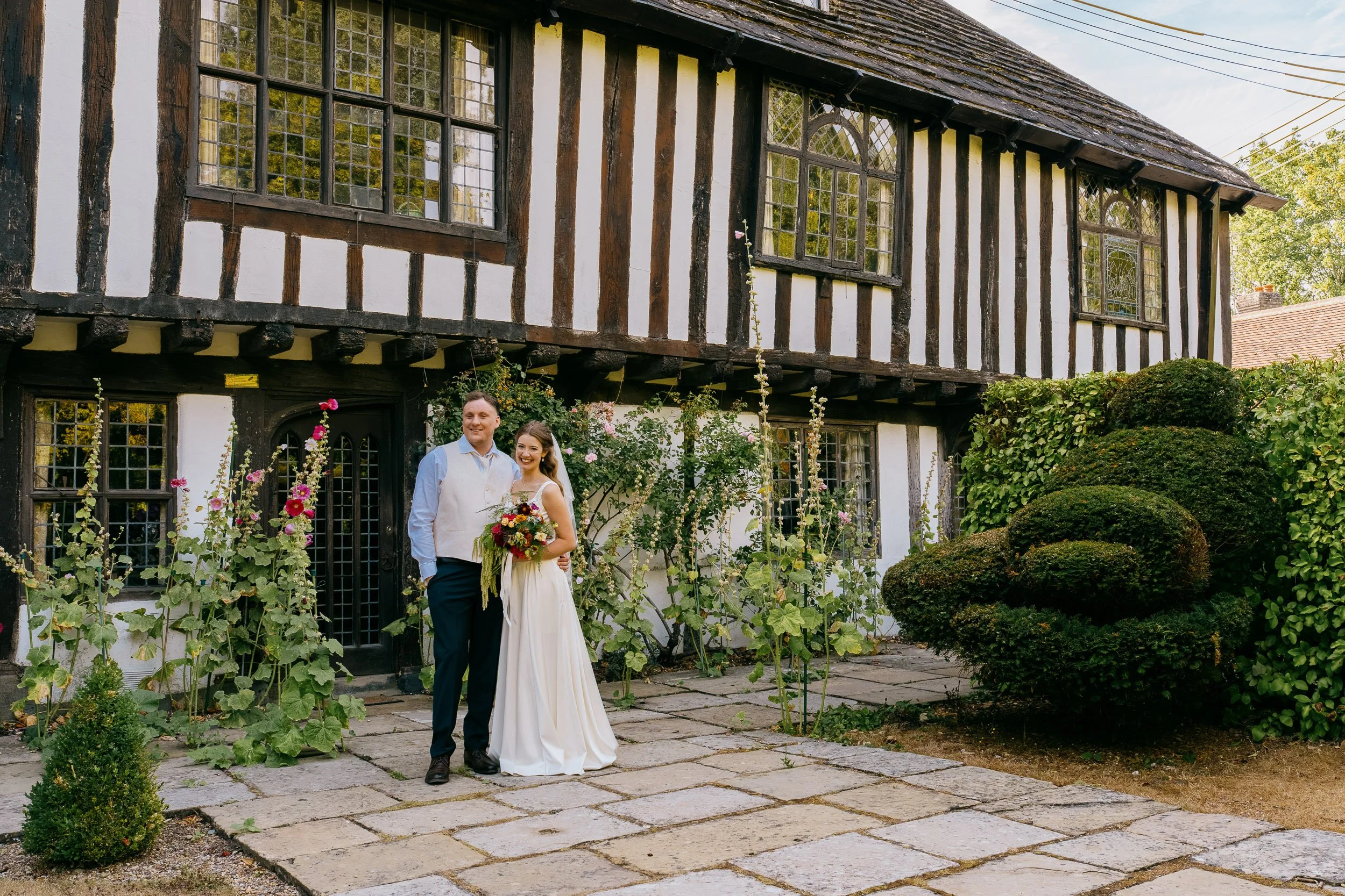 A happy couple, a man and a woman, dressed in wedding attire, standing in front of a historic half-timbered house with large leaded glass windows and surrounded by lush garden plants and flowers.