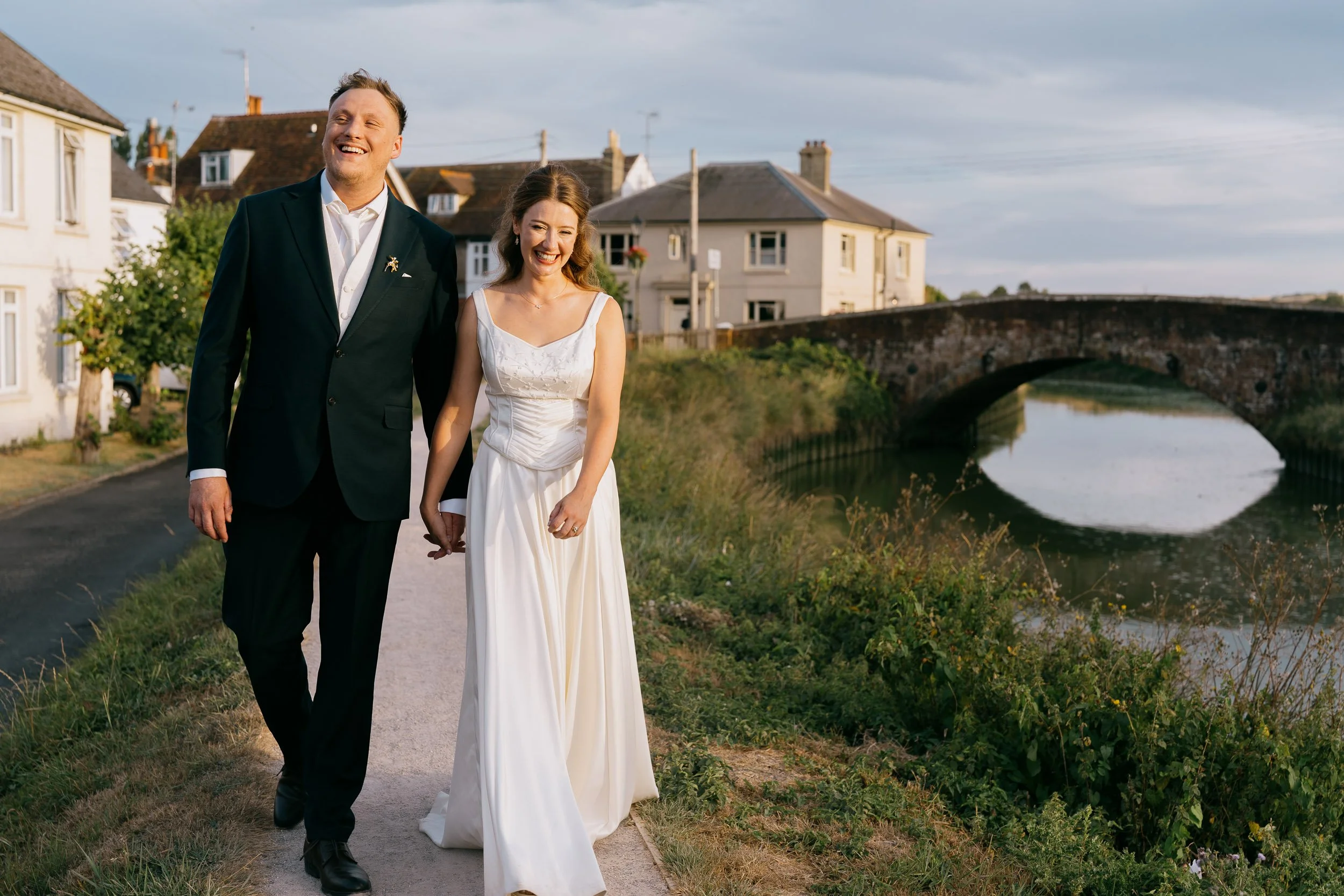 A smiling couple, dressed in wedding attire, walking hand in hand outdoors near a river and bridge with houses in the background.