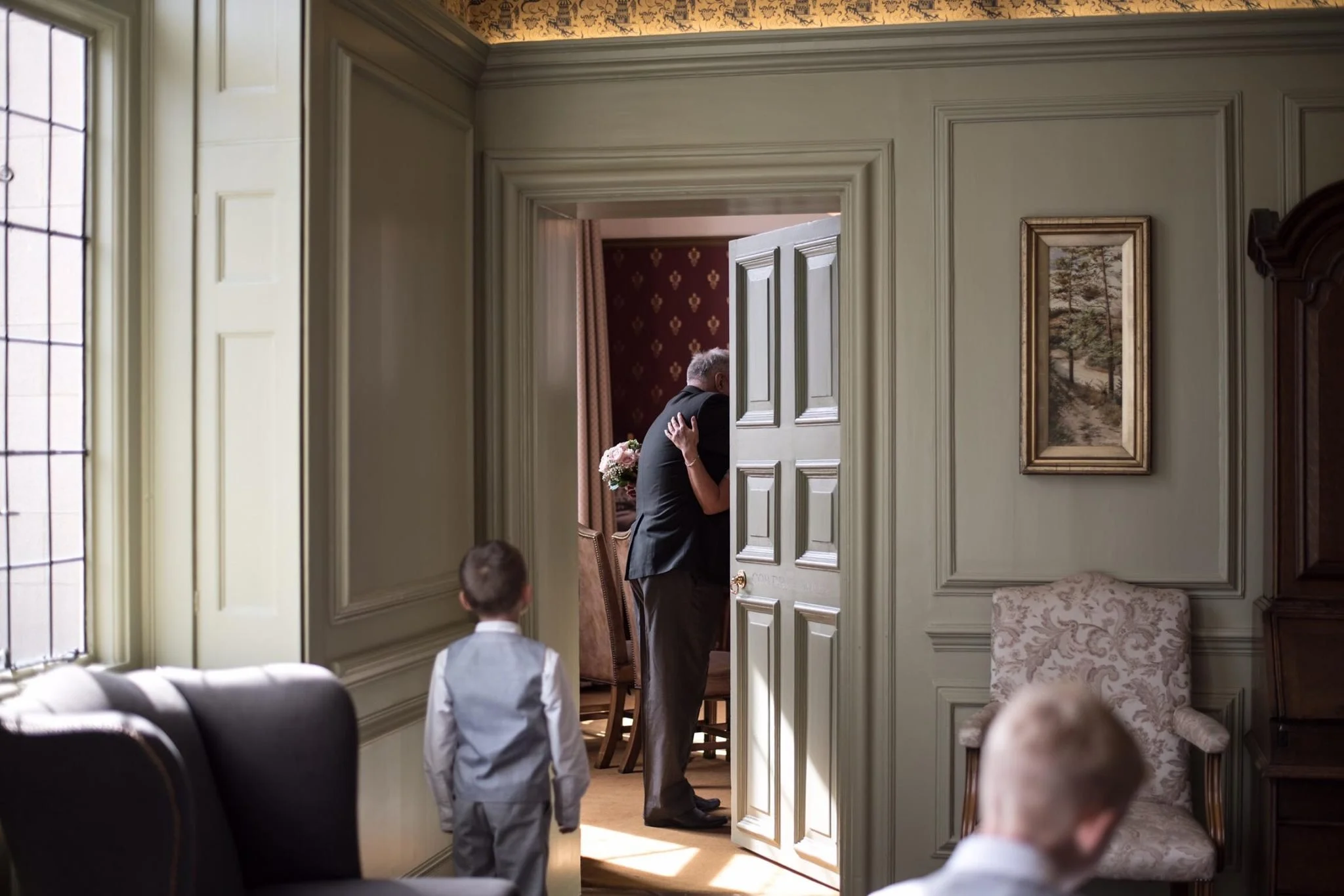 A man and woman embrace in a doorway, holding a bouquet of flowers, while two children in formal clothing look on in a room with upholstered chairs and framed artwork.