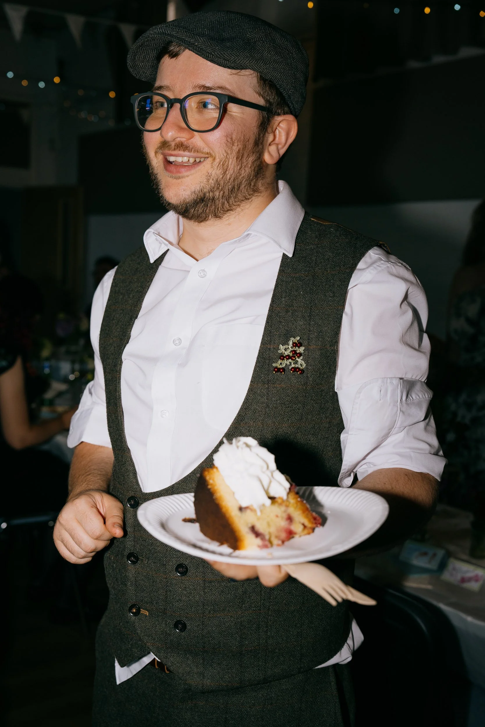 A man wearing glasses, a hat, and a vest holding a plate with a slice of cake.