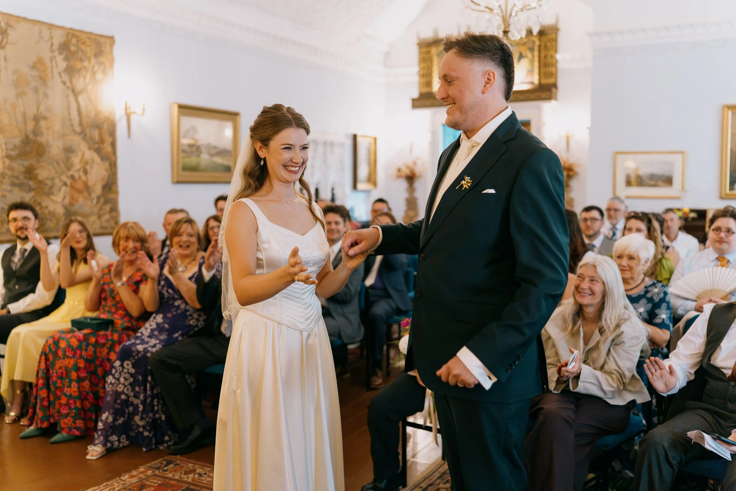 Bride and groom exchanging rings during wedding ceremony, guests seated and clapping in background, elegant decorated indoor setting.