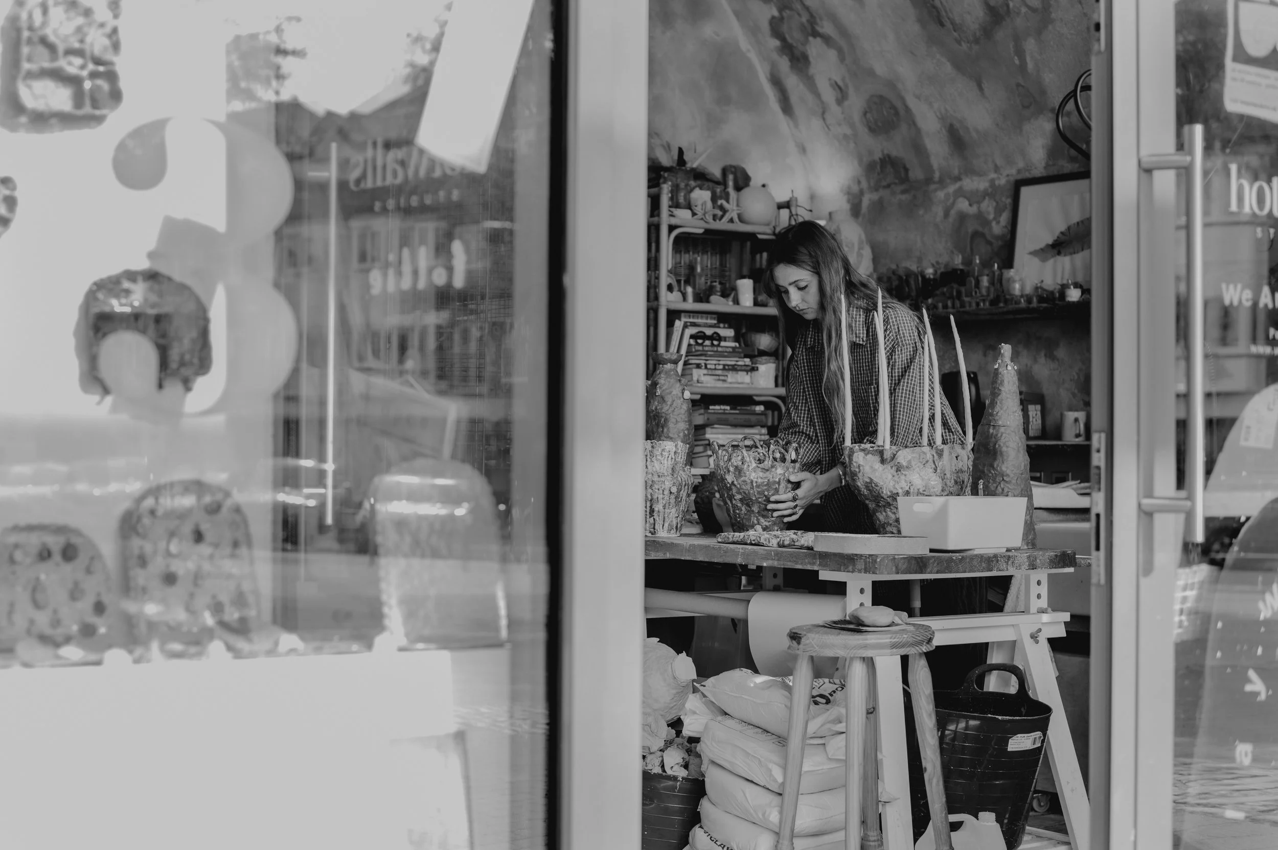 A woman working at her pottery studio, arranging clay sculptures on her table, seen through a glass window.