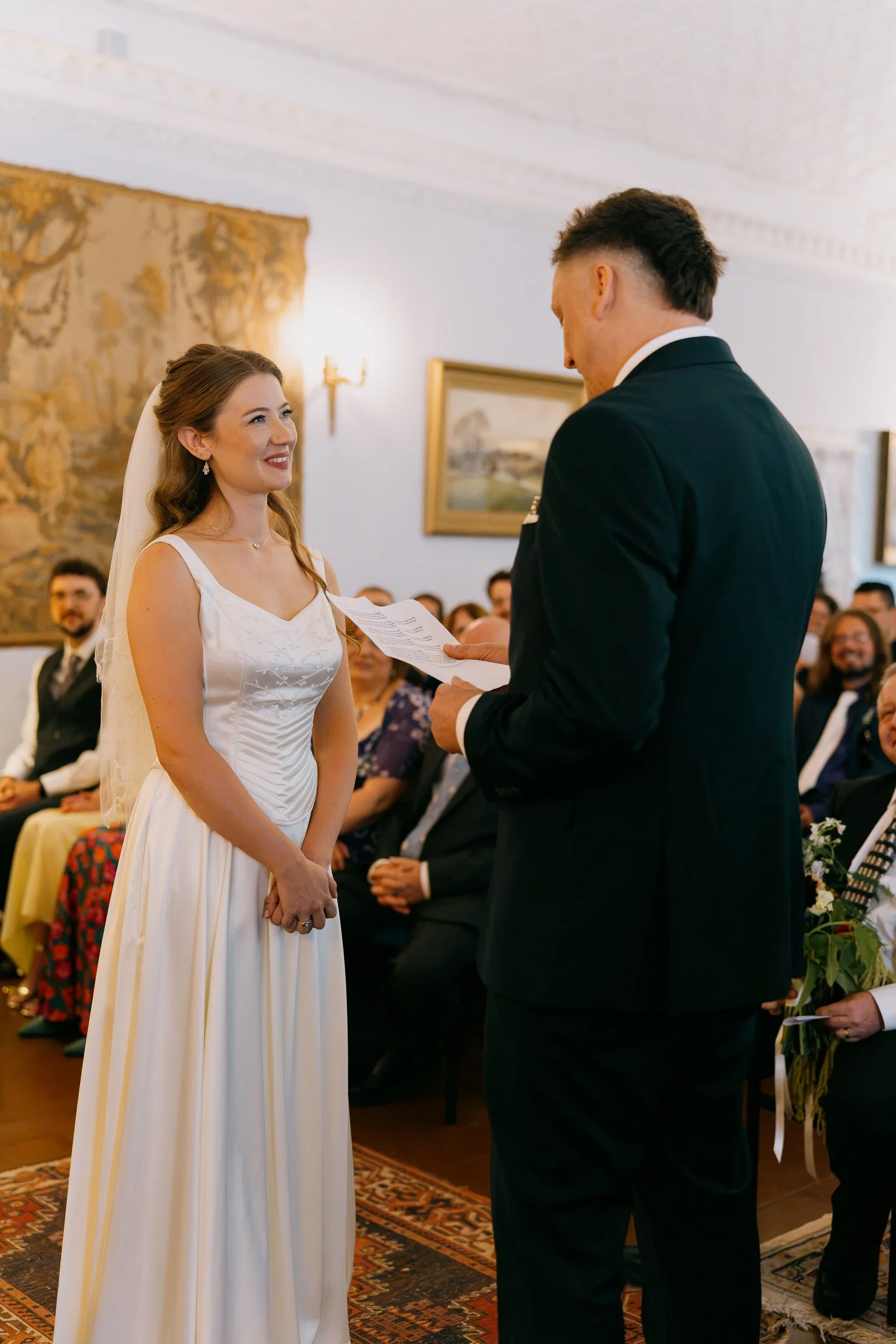 A bride and groom exchanging vows during their wedding ceremony in a decorated room with guests seated in the background.