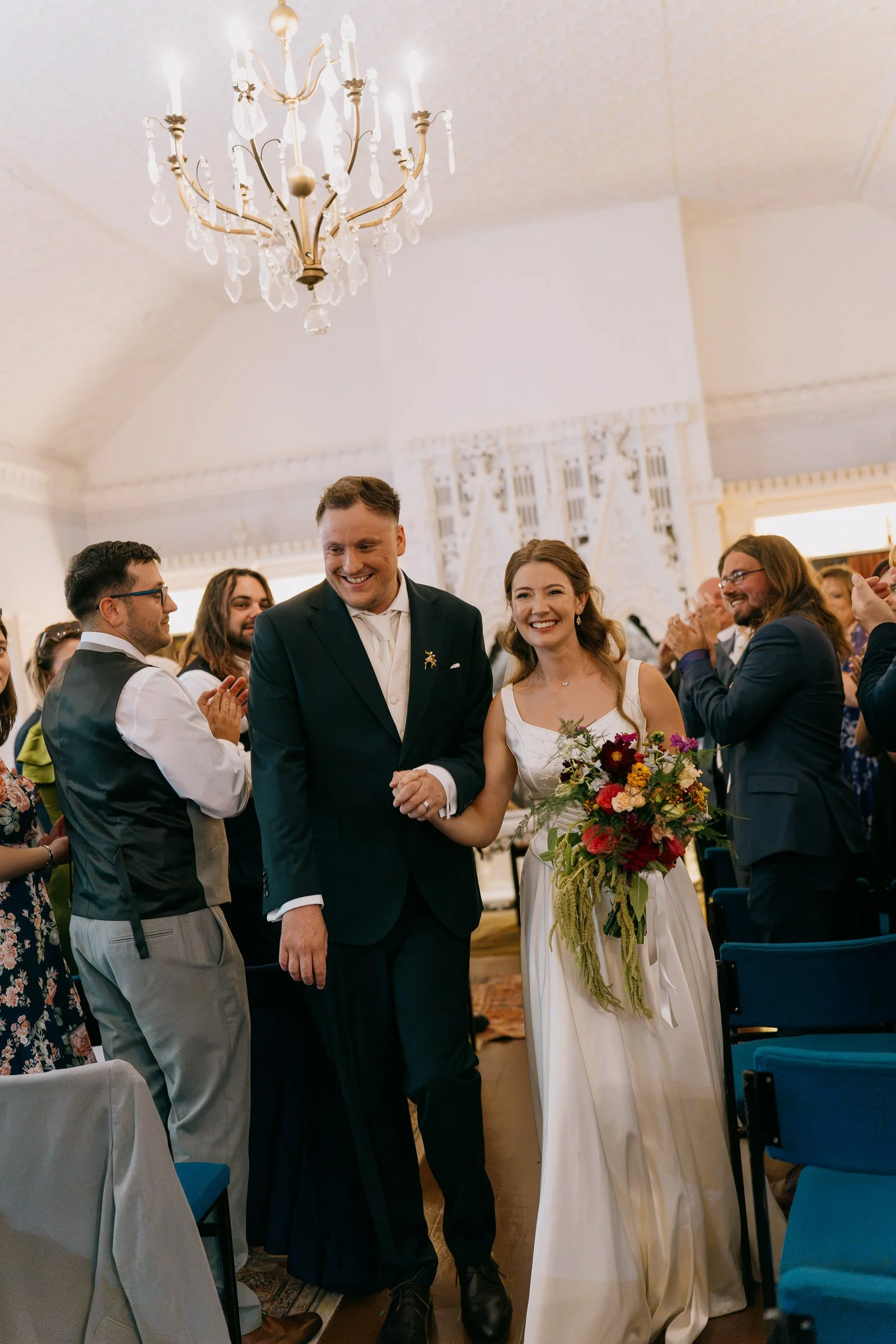 A wedding ceremony with a bride and groom holding hands smiling, surrounded by guests in a decorated room with a chandelier overhead.