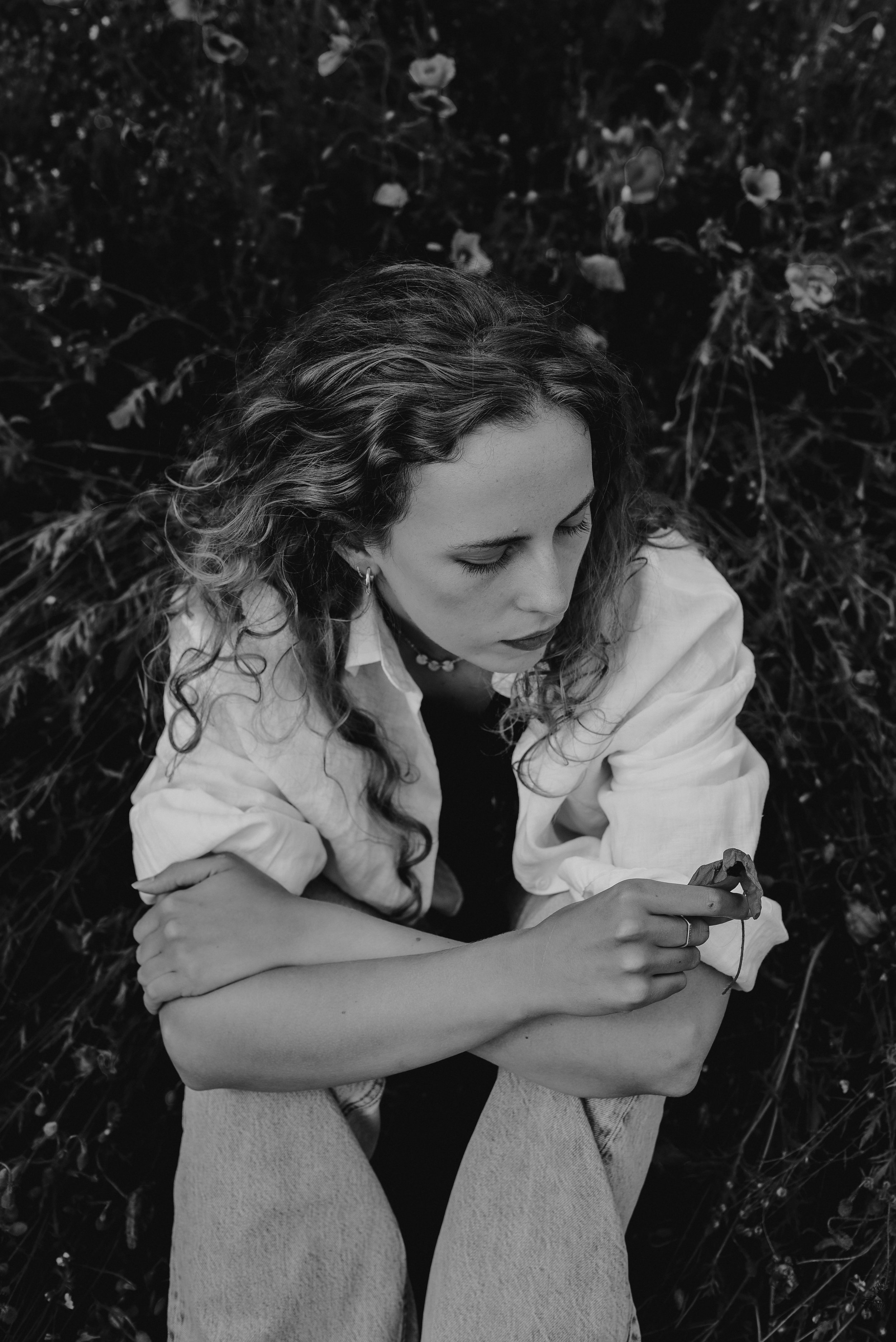 A woman with curly hair sitting outdoors on grass, looking down at a flower in her hand, in black and white.