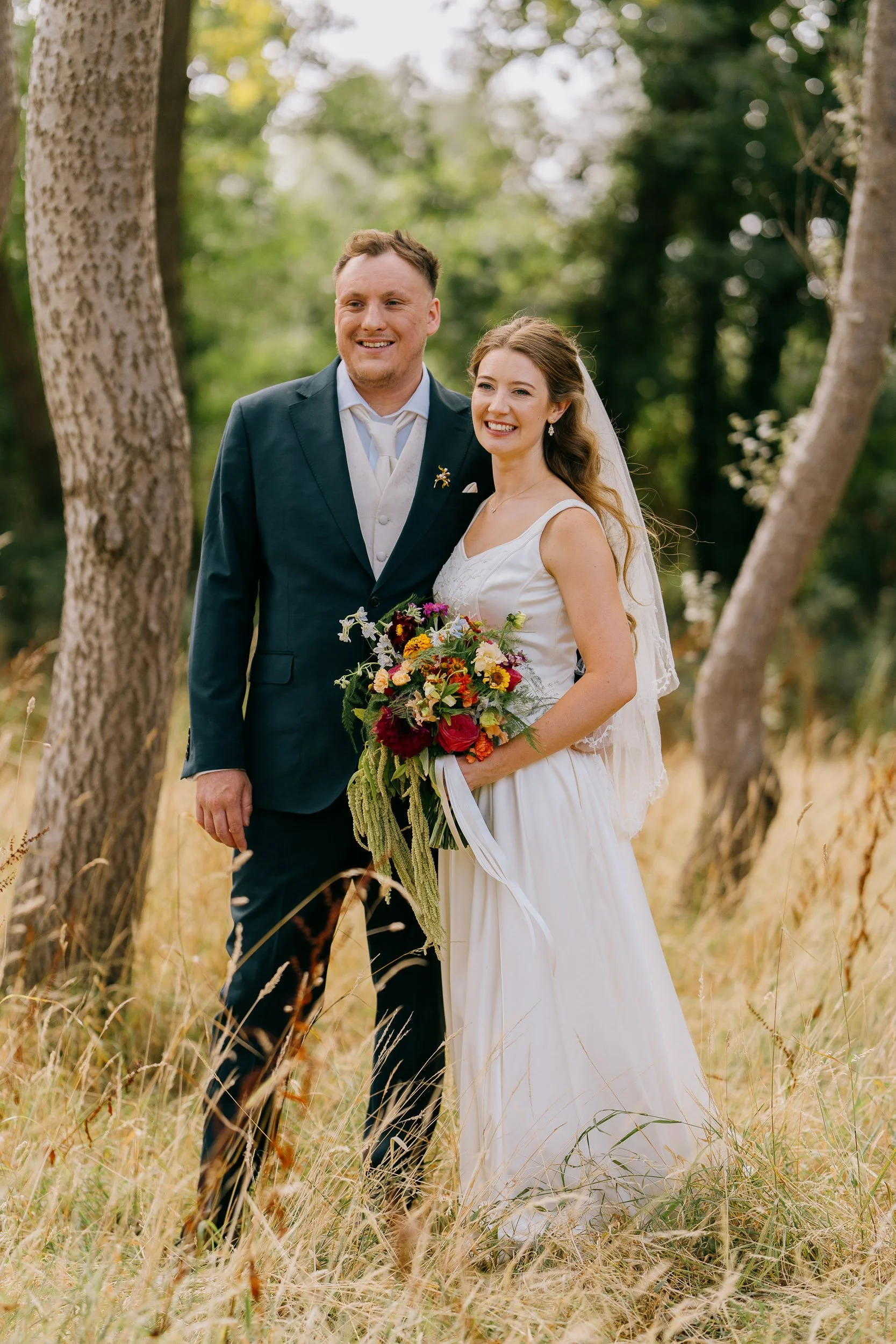 A bride and groom standing in a wooded outdoor setting, smiling and holding a bouquet of flowers.