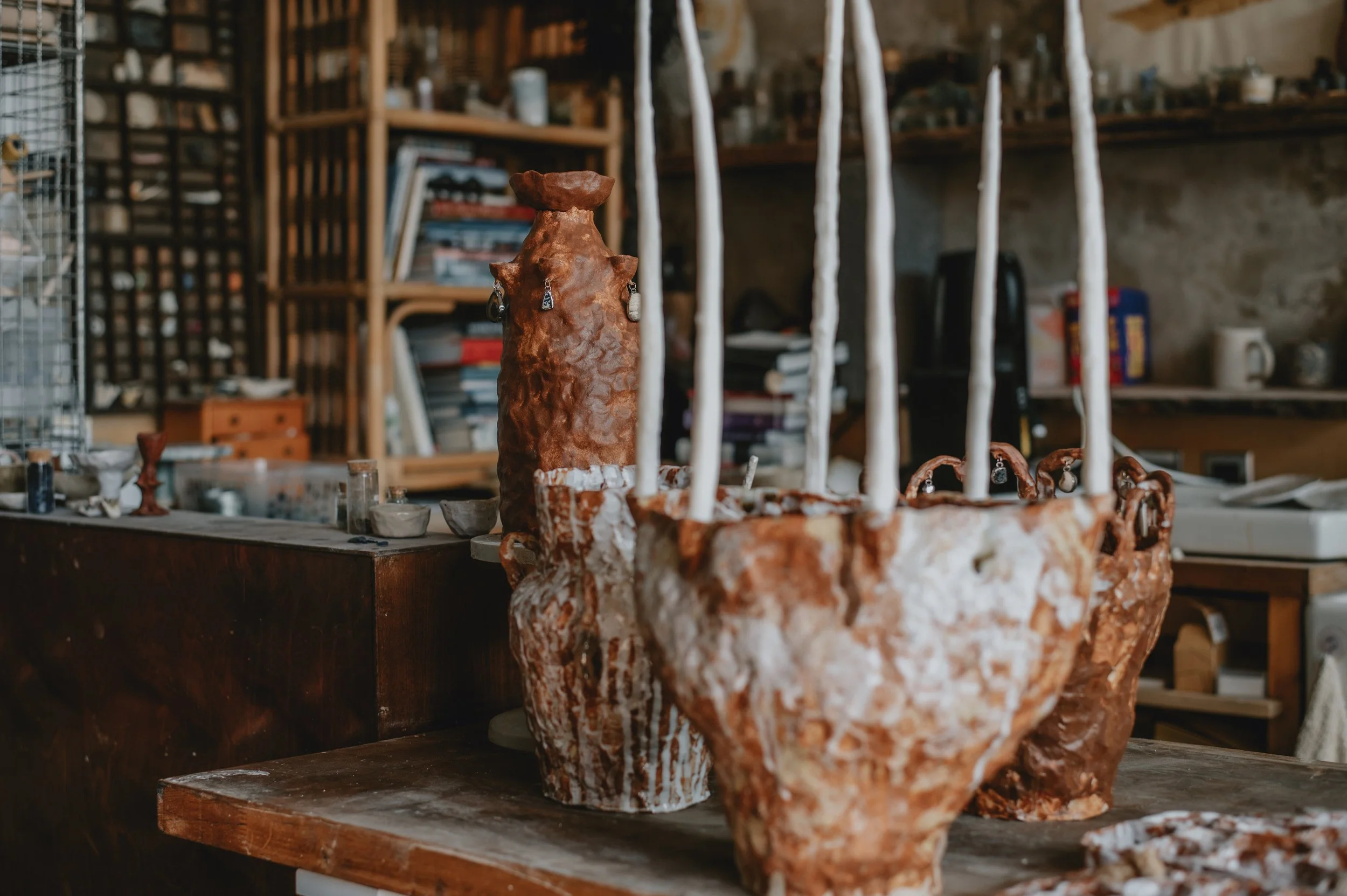 A rustic workshop with a reddish-brown textured clay sculpture of a dog with large ears and a small head, surrounded by various art supplies and books on shelves in the background.