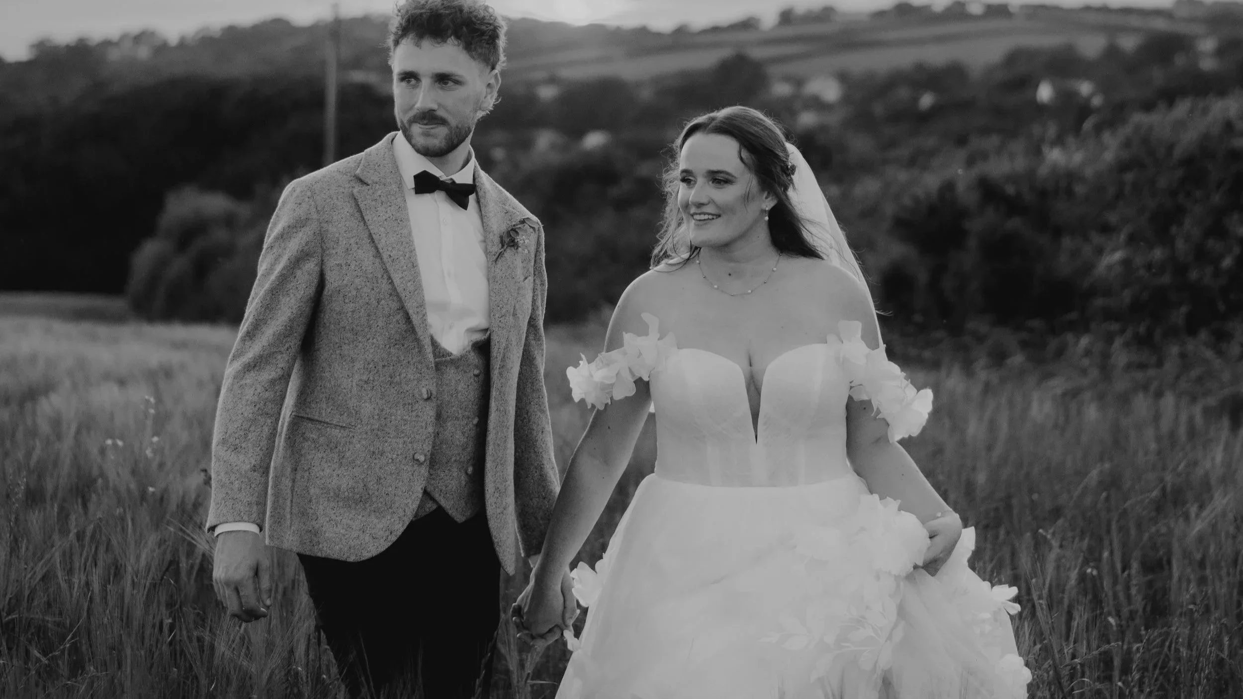 A black and white photo of a bride and groom holding hands in a field, smiling, with hills and trees in the background.