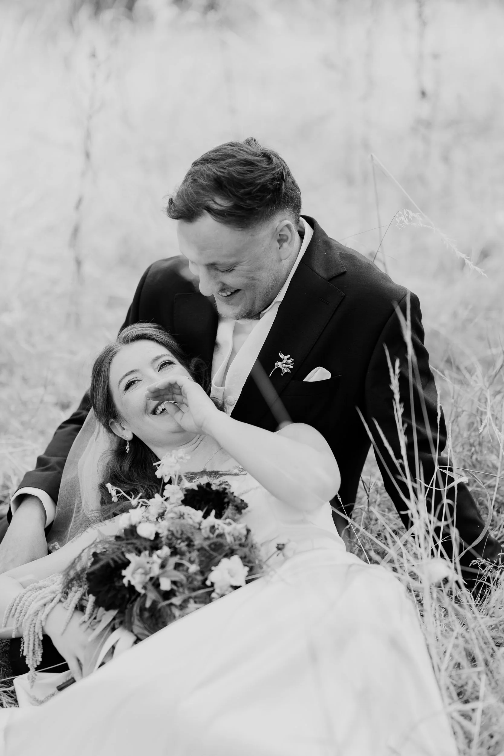 A black and white photo of a joyful couple lying on the grass outdoors, with the woman holding a bouquet of flowers.